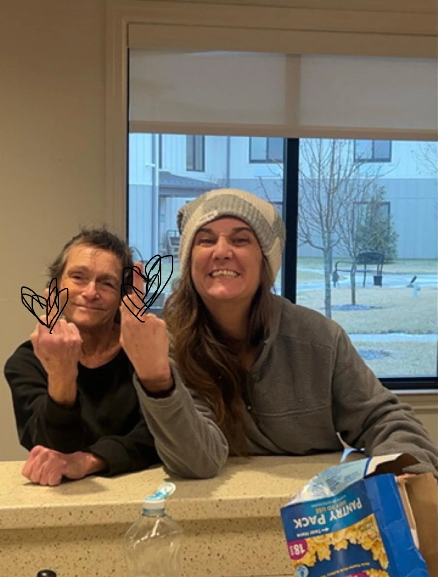 Two people sitting at a kitchen counter with a box of microwave popcorn in the foreground. One person is making a fist with a smile, the other is smiling with a beanie hat. A window with a view of trees and buildings is in the background.