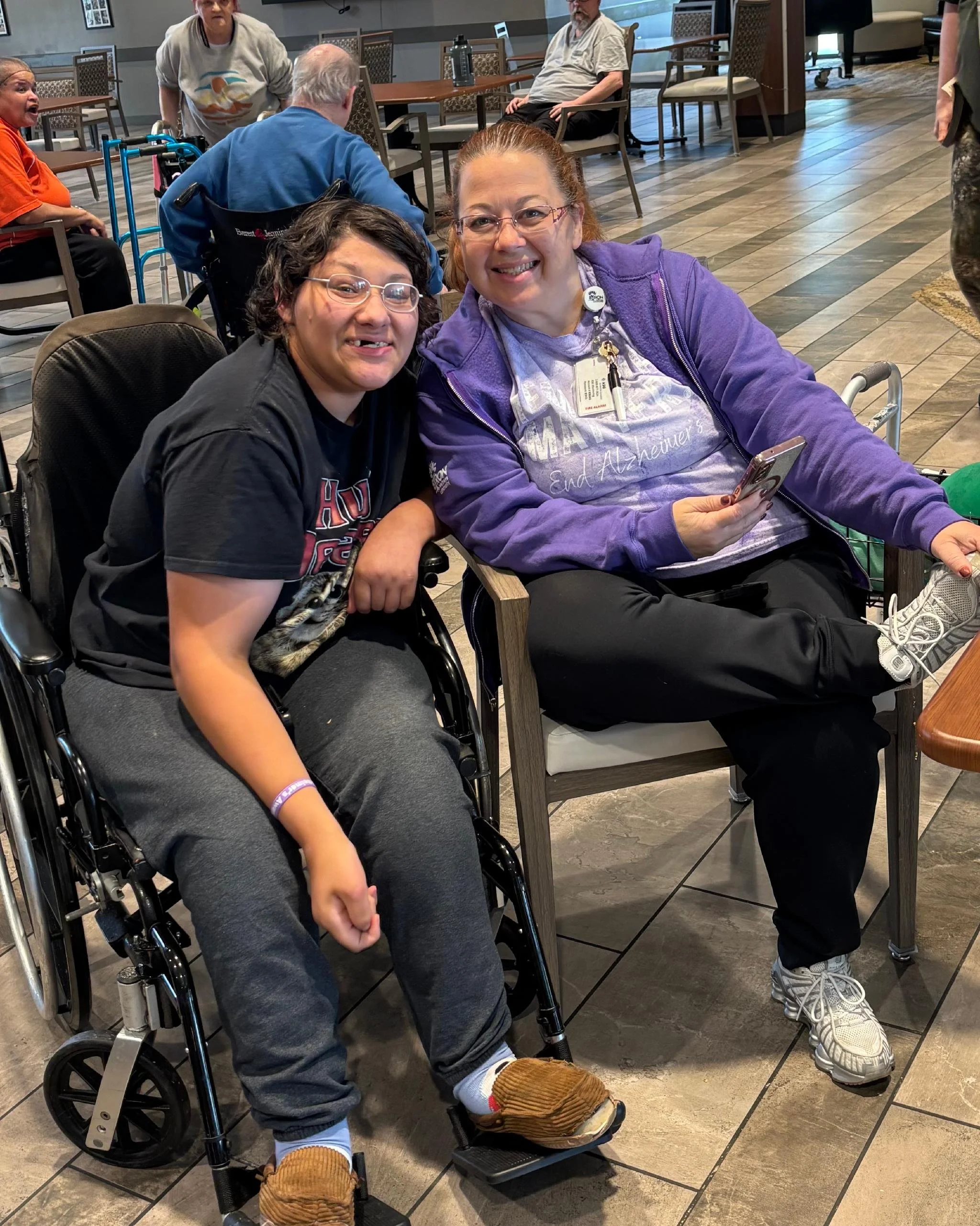 Two women with disabilities pose for a photo inside a dining area, smiling. Behind them are several people, some seated at tables and some standing, in a room with tiled floors and chairs.