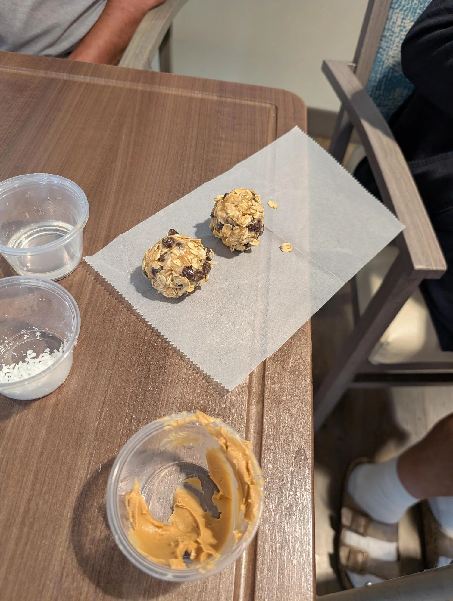 Two oatmeal balls with raisins on a white paper on a wooden table next to small plastic containers, one containing remaining peanut butter.