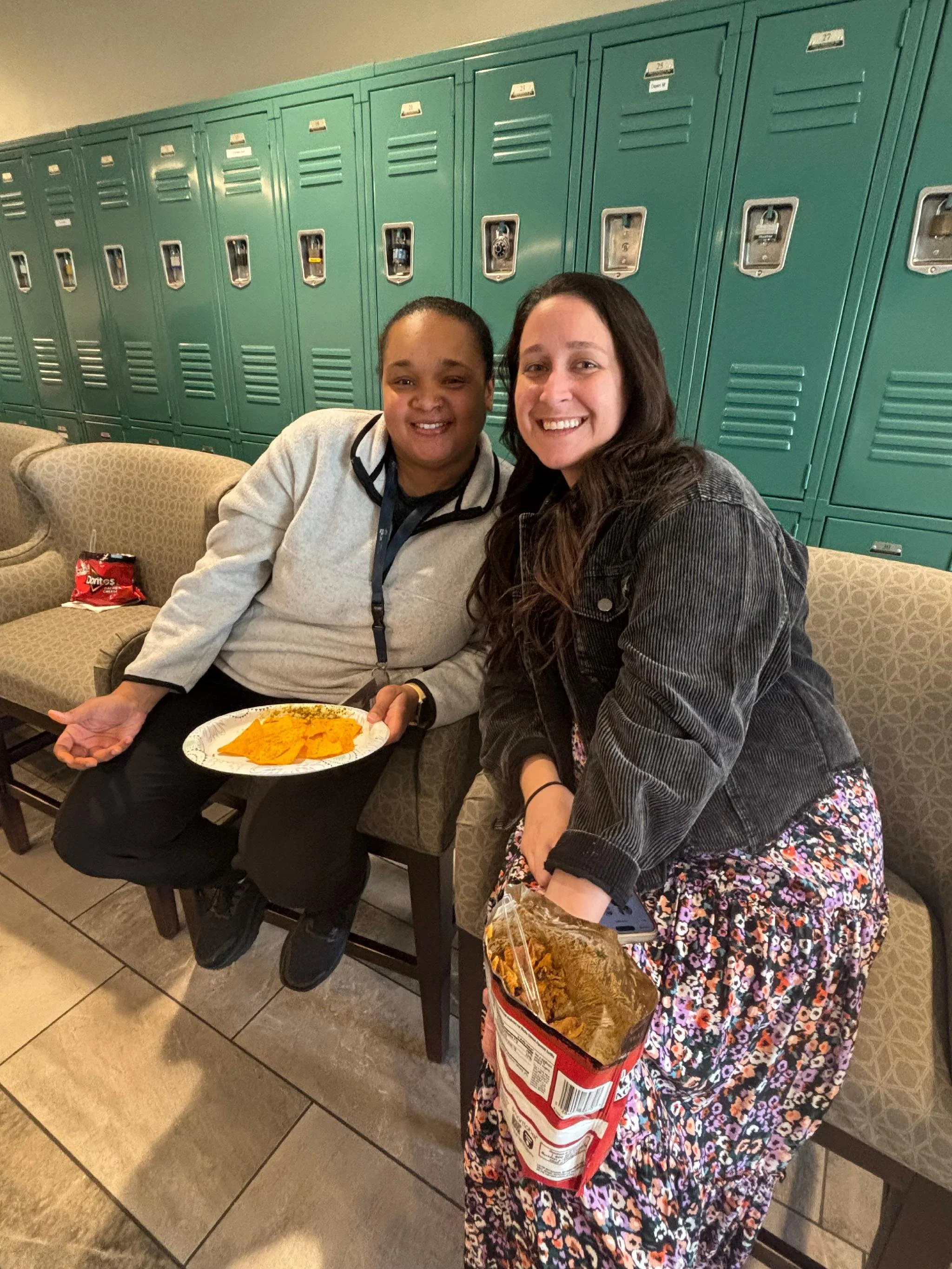 Two women sitting on a bench in front of teal lockers in a school hallway, smiling at the camera. One woman is holding a plate of nachos, and the other is holding a bag of chips.
