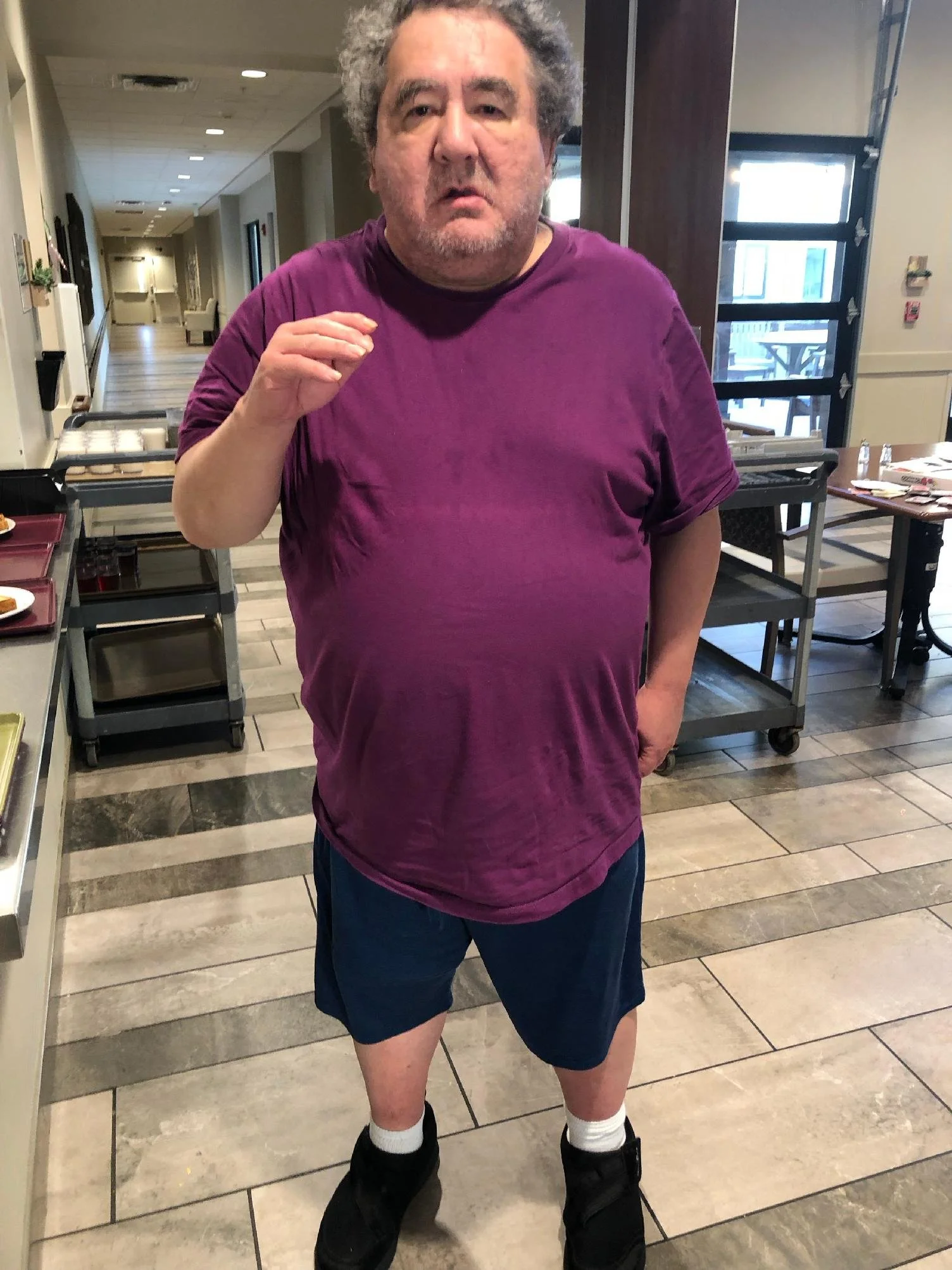 A man is standing in a restaurant, wearing a purple t-shirt, black shorts, white socks, and black shoes. The man appears to be looking at the camera with a neutral expression.