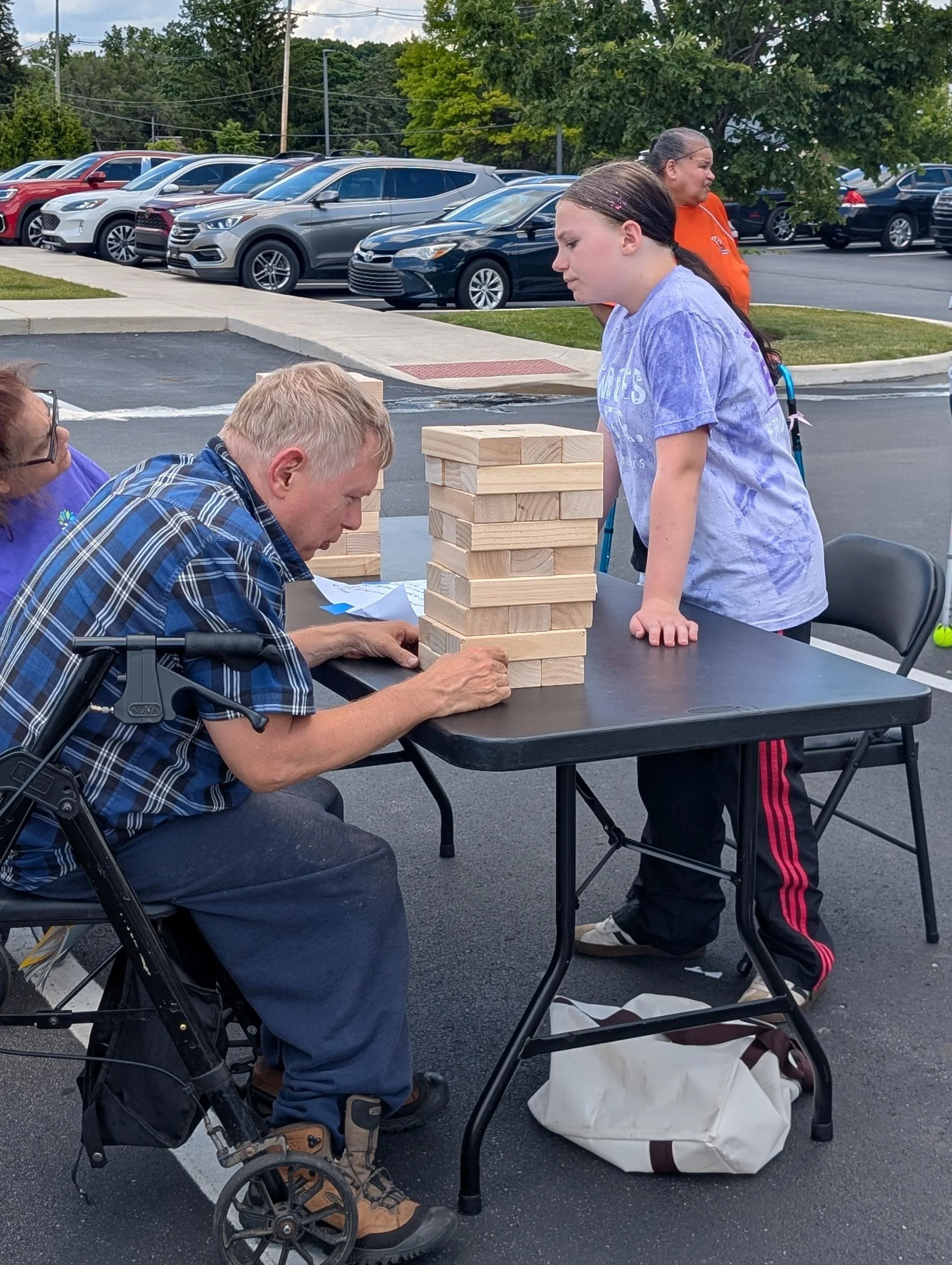 People playing Jenga outdoors in a parking lot. A young girl is standing, while an elderly man in a wheelchair and a woman sit nearby.
