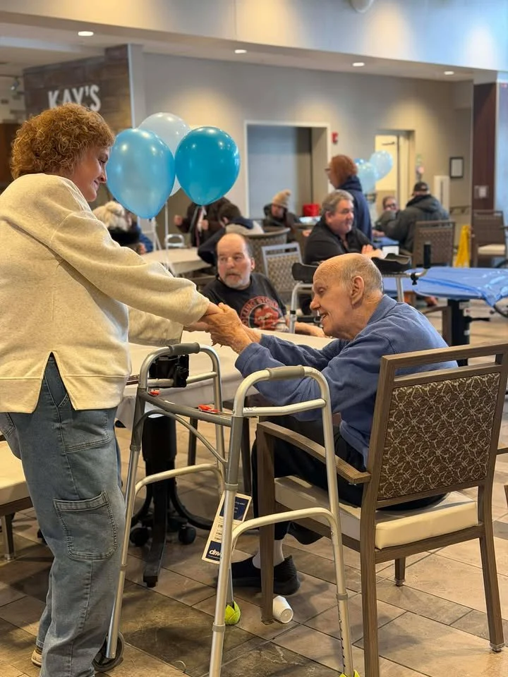 An elderly man in a wheelchair shaking hands with a woman in a beige sweater at a social gathering in a dining area of what seems to be a senior living center, decorated with blue balloons and other seniors dining in the background.
