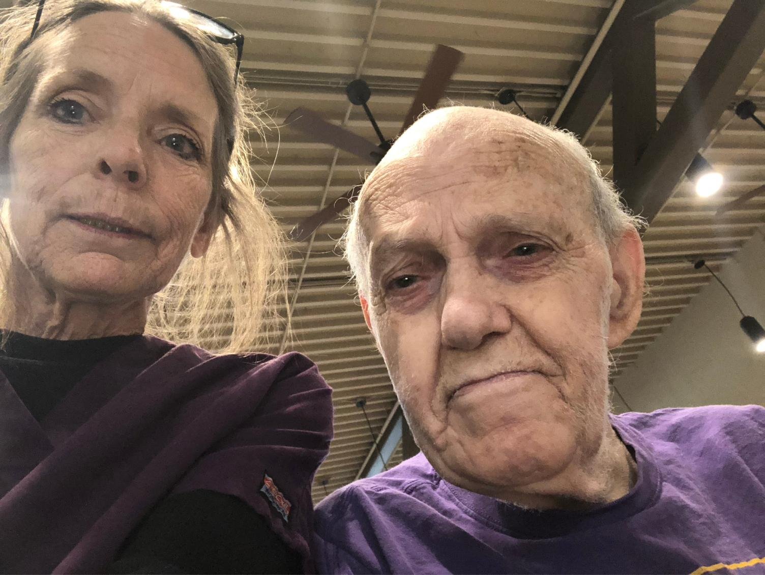 Close-up selfie of an elderly man and woman inside a building with a modern ceiling design and hanging lights