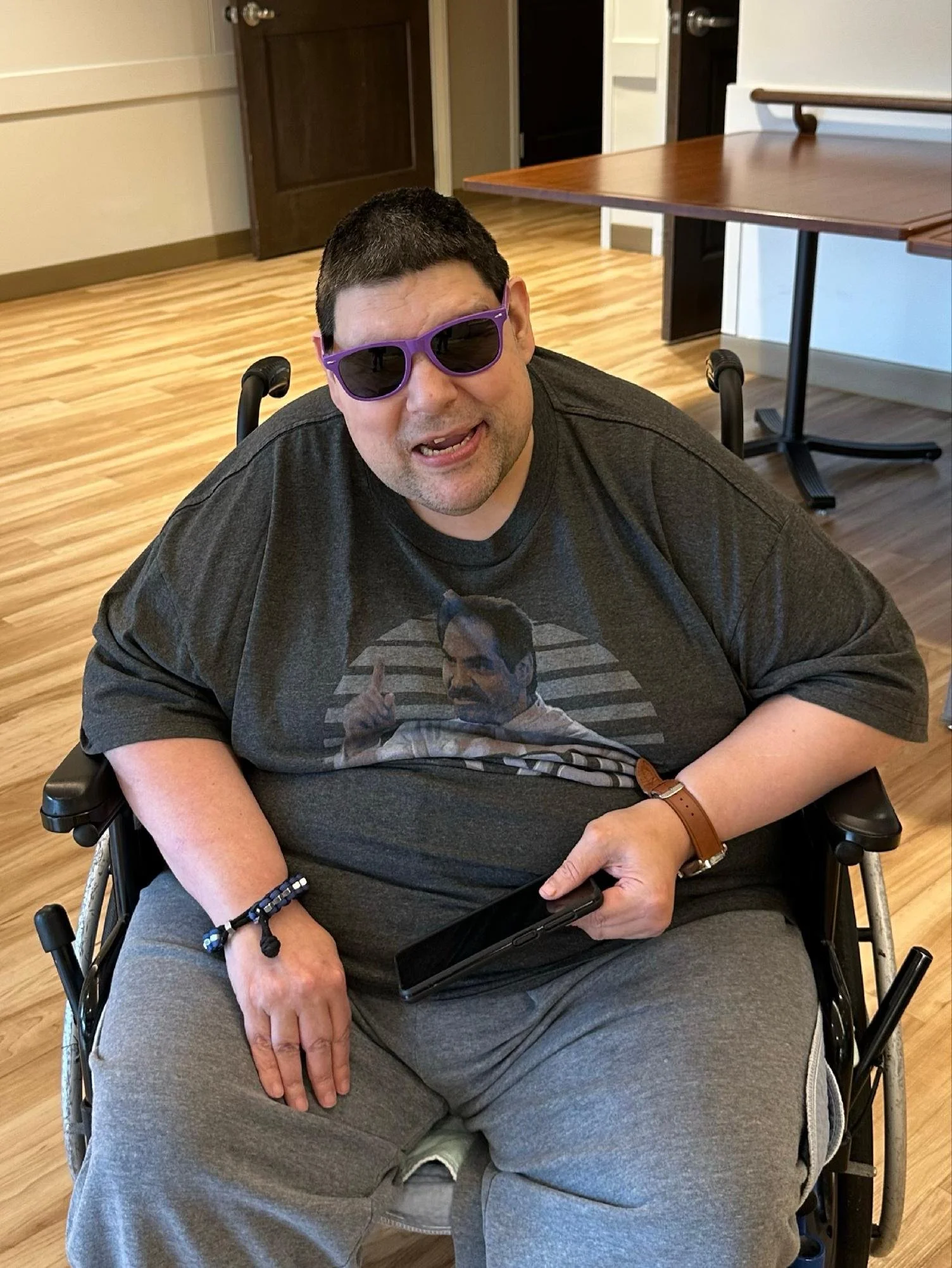 A man in a wheelchair wearing purple sunglasses and a dark gray t-shirt with a graphic of a man pointing. He has a short haircut, a brown wristwatch, and wristbands, and is holding a phone in his hand. The background shows a wooden floor, a brown doo