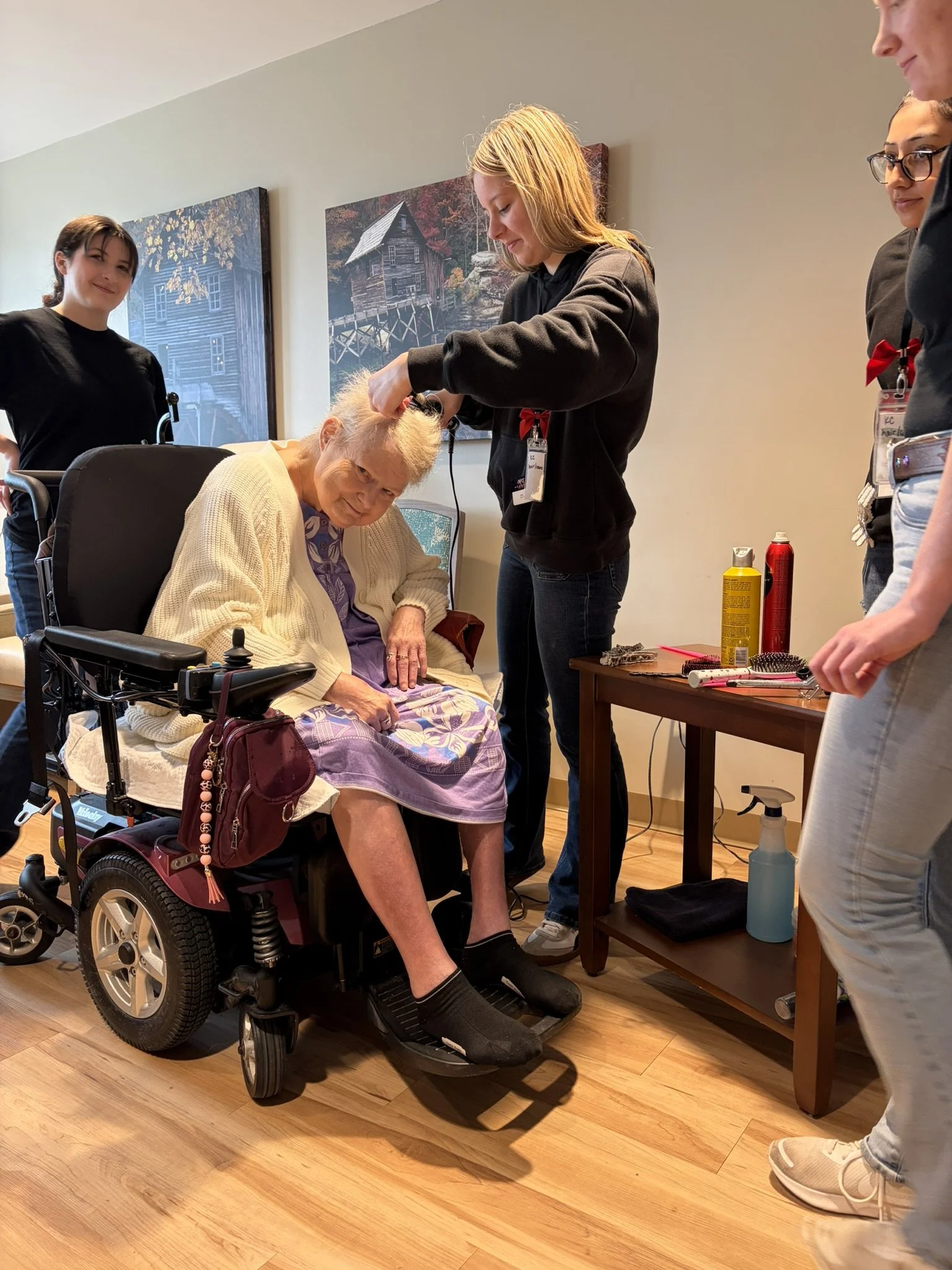 A woman with white hair sitting in a motorized wheelchair in a room with wooden floors. She is receiving a haircut from a young woman with blonde hair. Several other people are standing around, observing the haircut. The room has two large paintings 