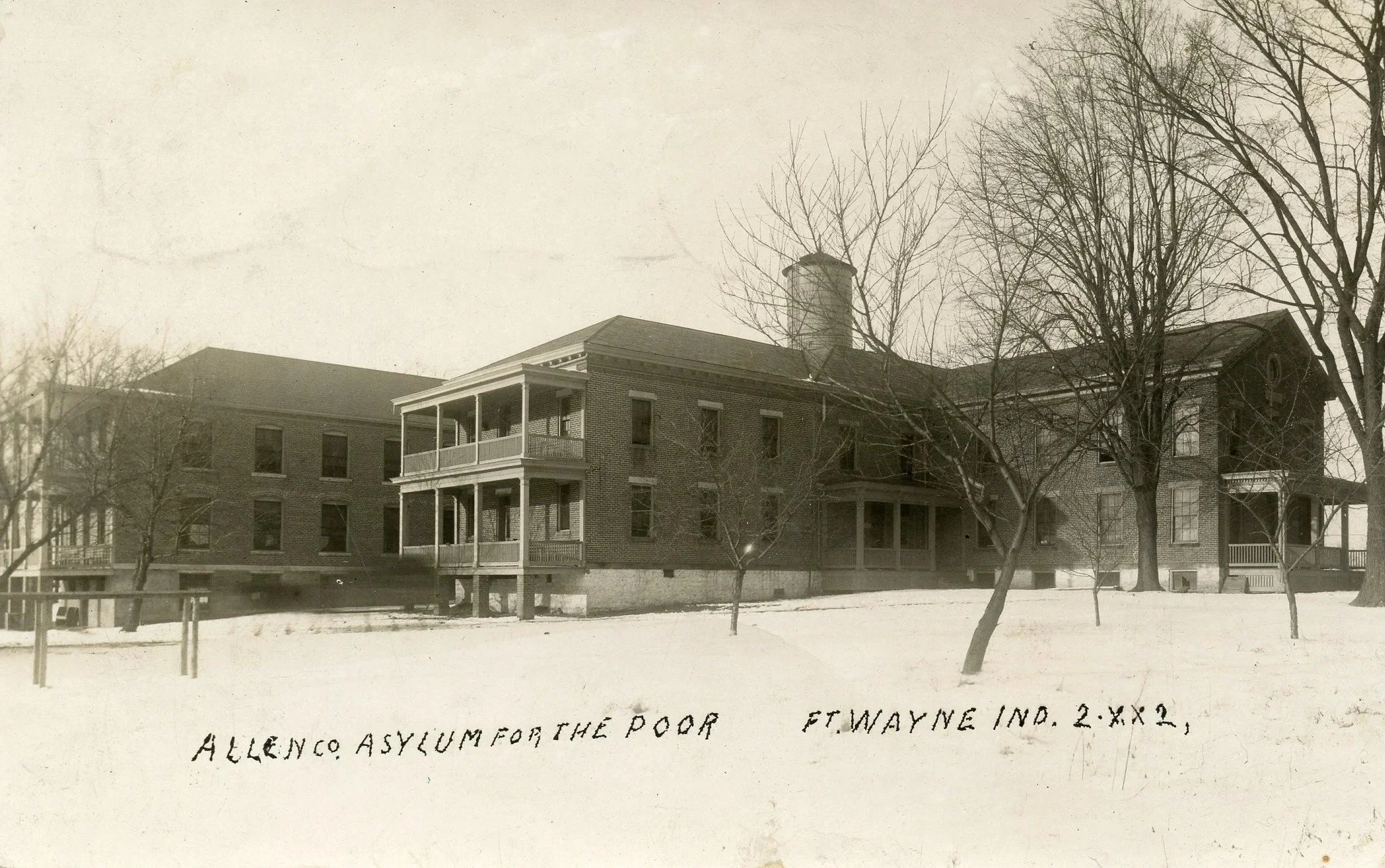 A black and white photograph of a large, three-story brick building with a prominent chimney, surrounded by leafless trees. The building has multiple balconies and windows. At the bottom, handwritten text reads: "ALLEN CO. ASYLUM FOR THE POOR FT. WAYNE IND. 2 XX 2."