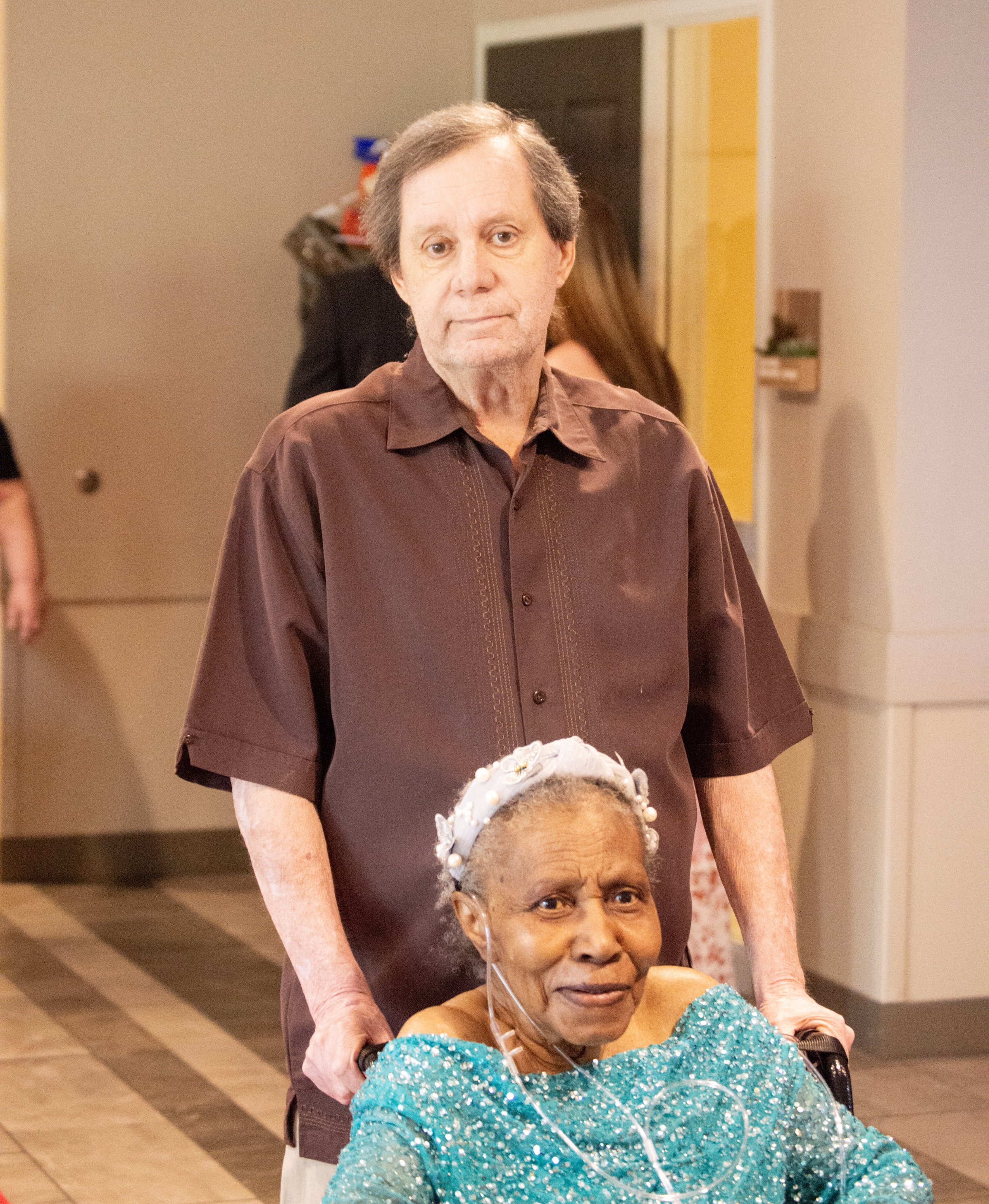 An elderly woman with a pearl-embellished gray headband sitting in a wheelchair, wearing a sparkly teal dress, with a man standing behind her, holding her shoulders, in an indoor setting.
