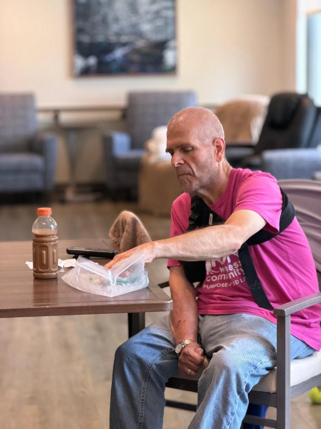An elderly man with a shaved head wearing a pink shirt with black straps, sitting in a chair at a table, reaching into a plastic bag. On the table, there is a bottle of chocolate milk and a remote control.