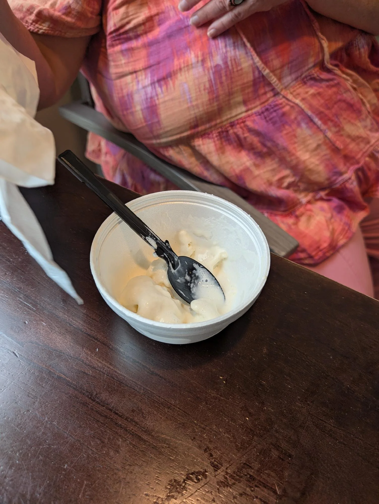 A plastic cup of vanilla ice cream with a black plastic spoon on top, sitting on a dark wooden table. Part of a person's body wearing a colorful, patterned shirt is visible in the background.