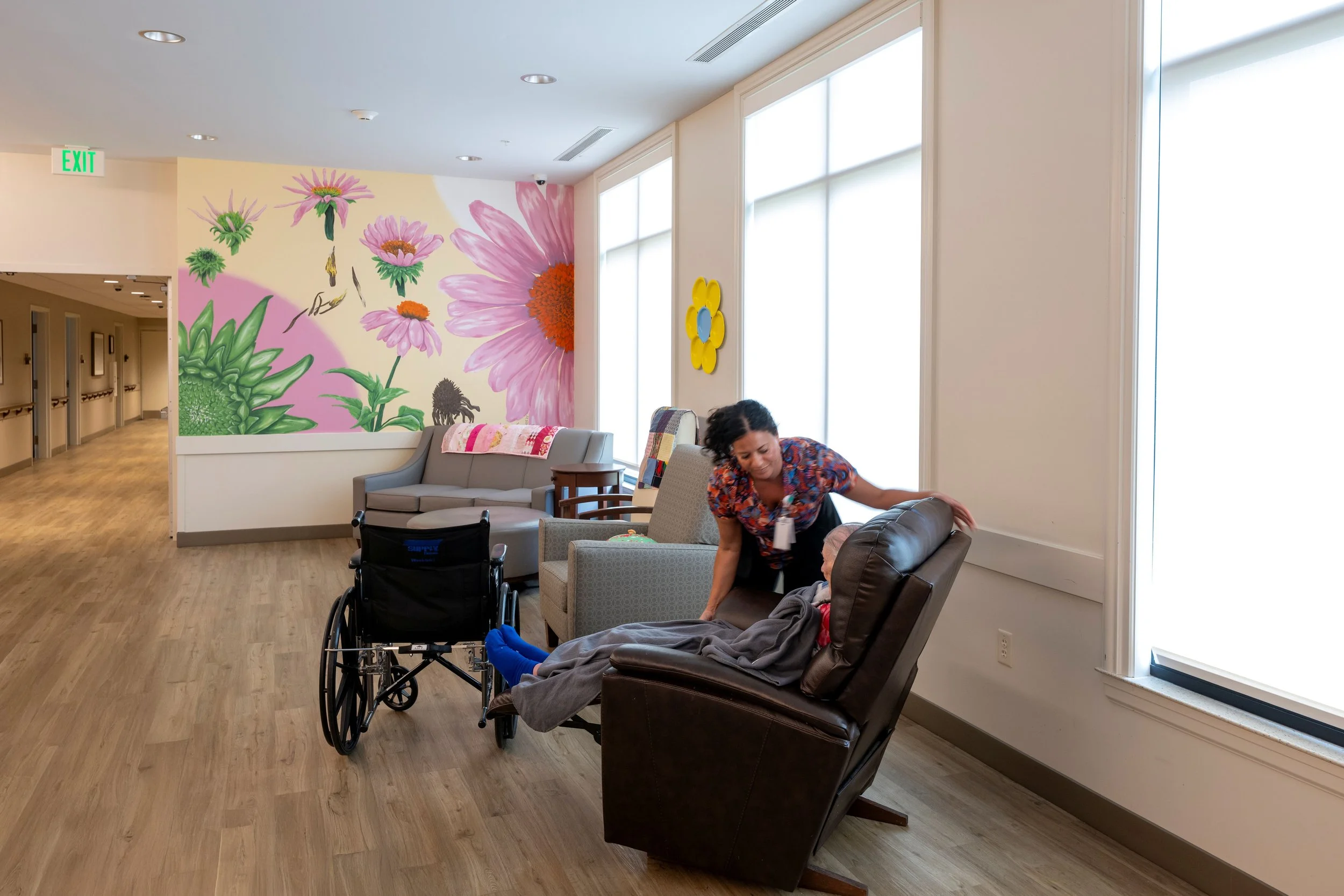 A woman with dark curly hair sitting on a beige armchair interacting with a person in a wheelchair in a room with large windows and a colorful floral mural on the wall.