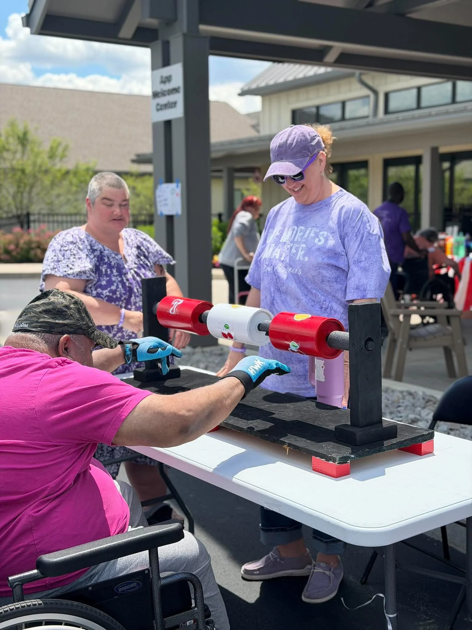 People at an outdoor event with a table, some holding mugs, and a shuffleboard game in front of a building with a sign that says 'Dog Wellness Center'.