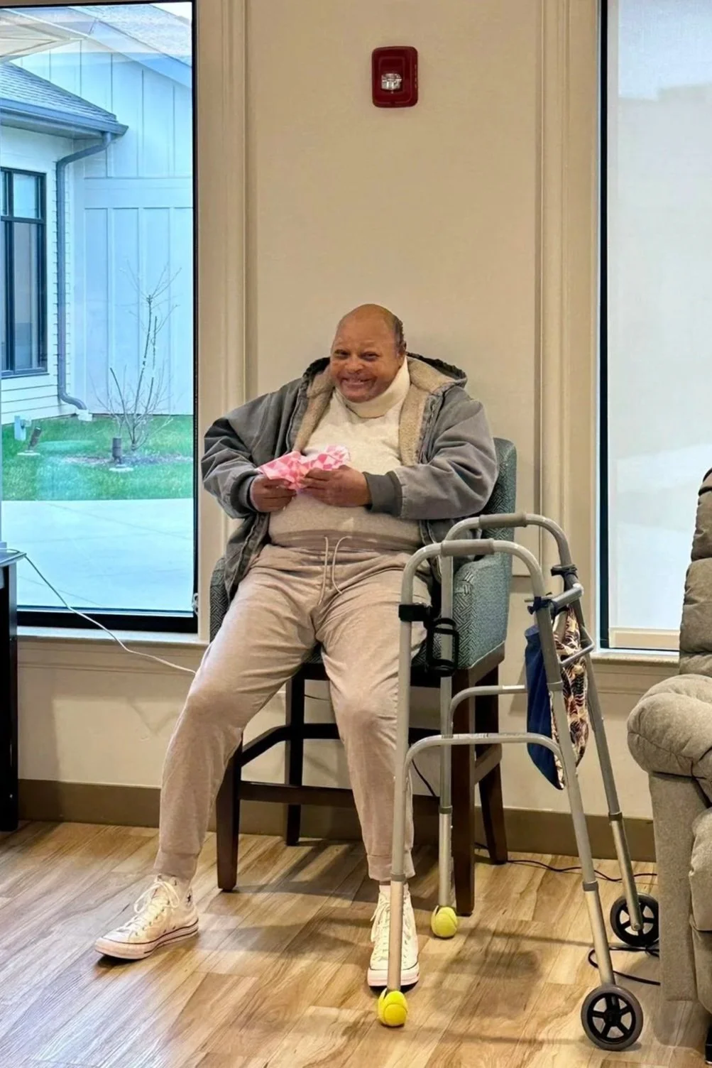 A man sitting on a high chair near a window, holding a wrapped gift, with a walker in front of him, smiling in an indoor setting.