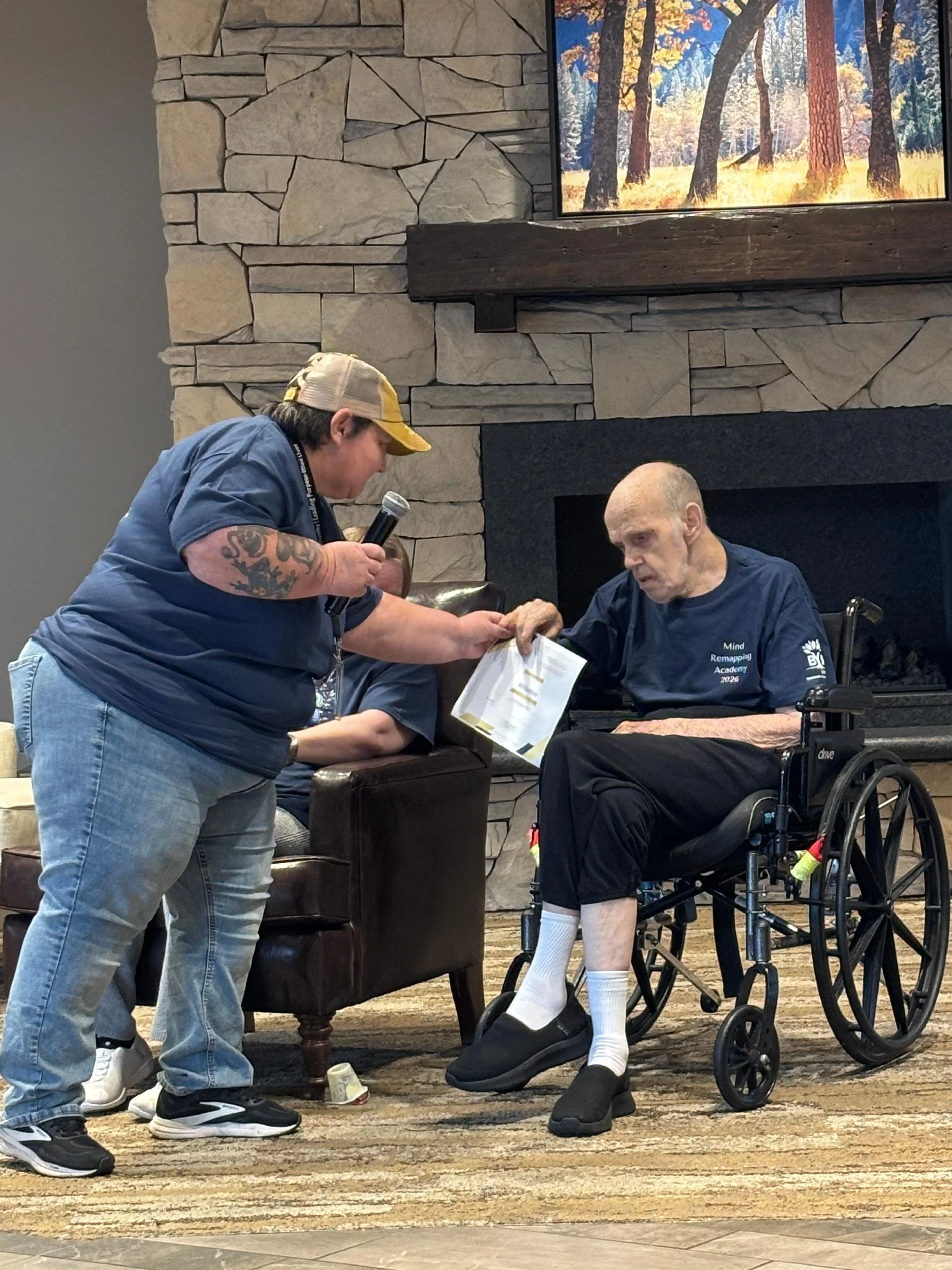 A woman with tattoos on her left arm wearing a baseball cap and blue t-shirt is handing something to an elderly man in a wheelchair. The elderly man has a bald head, wears a dark t-shirt with writing on it, white socks, and black shoes. They are indo
