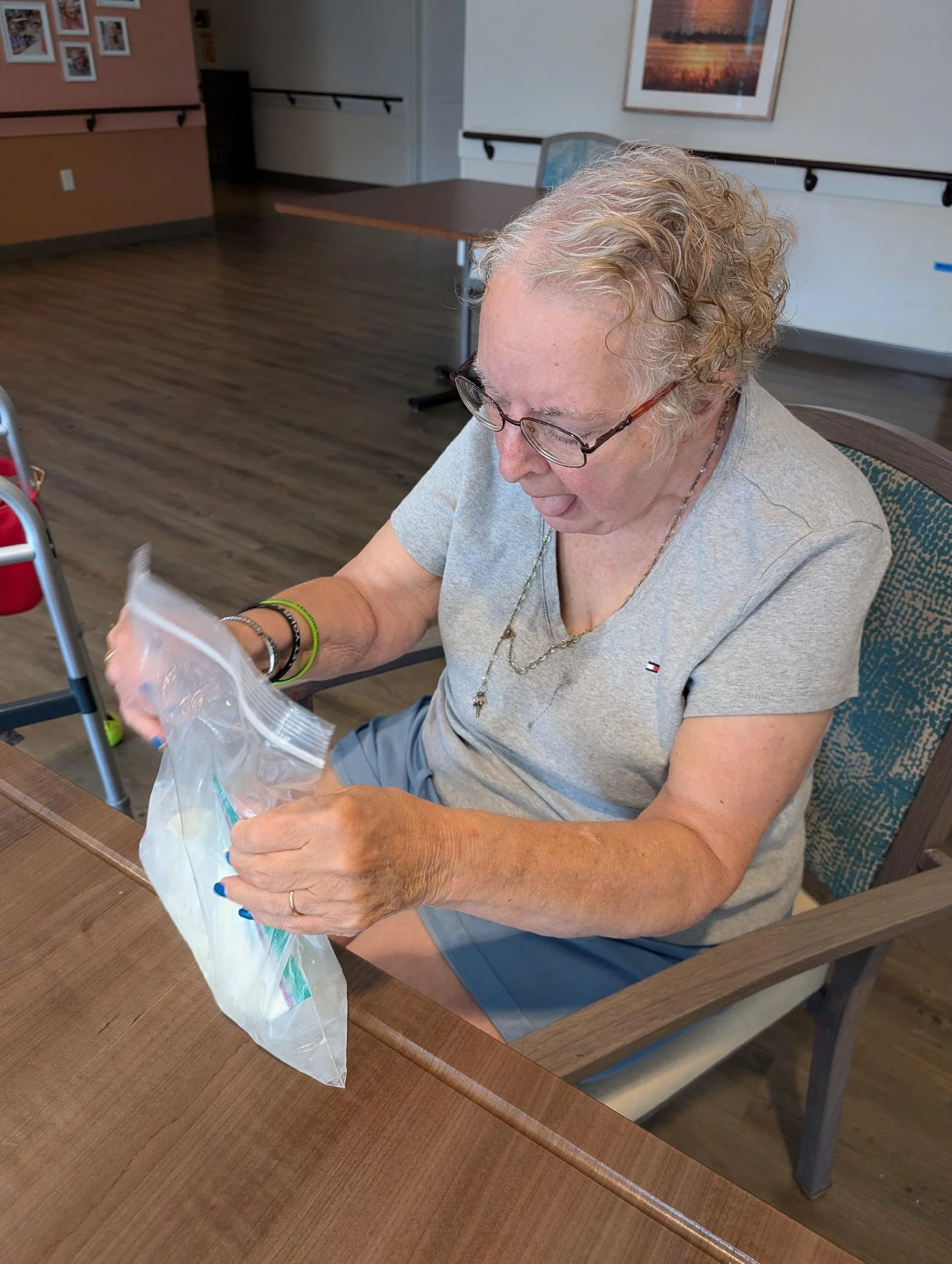 An elderly woman with short curly gray hair and glasses, sitting at a wooden table in a room, opening a clear plastic bag.