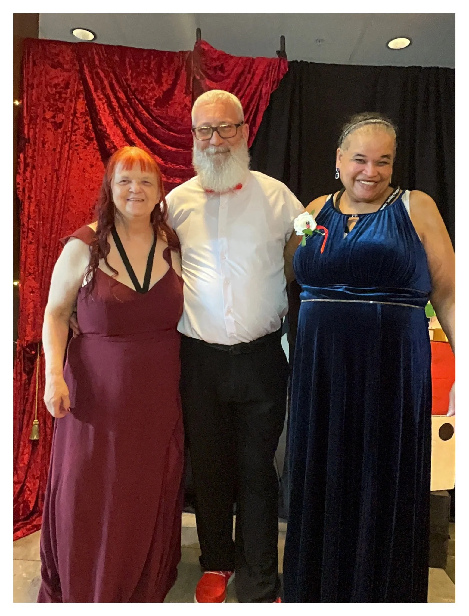 Three people standing in front of a red and black draped backdrop, dressed in formal attire, smiling for a photo.