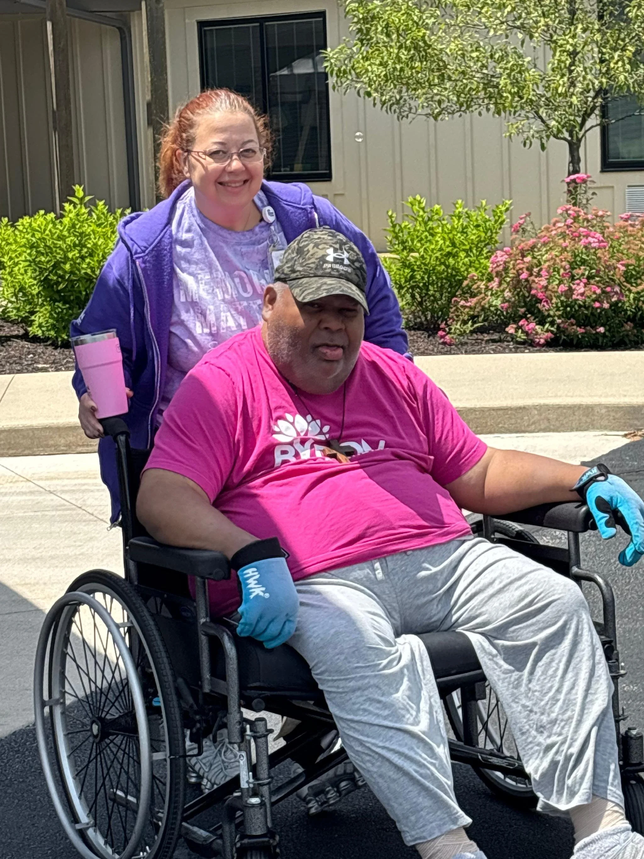 A woman with glasses and curly hair stands behind a man in a wheelchair. They are outdoors with bushes and a building in the background. The woman is smiling and wearing a purple jacket, and the man, dressed in a pink shirt and camouflage cap, is sti