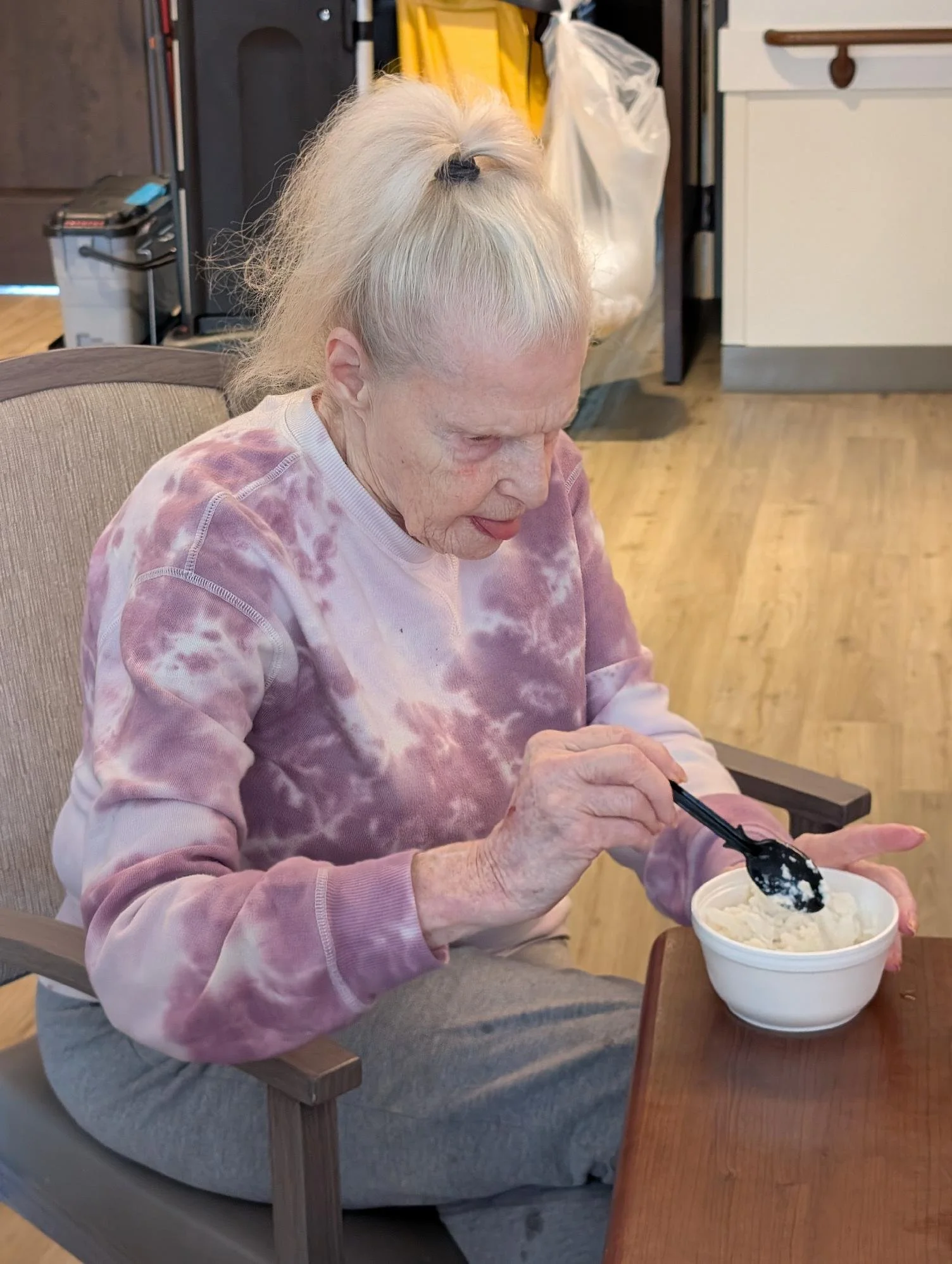 An elderly woman with long white hair tied in a high ponytail, wearing a pink and purple tie-dye sweatshirt, is sitting in a gray armchair, enjoying ice cream in a white bowl. She is using a black spoon with some ice cream on it, and there is a small