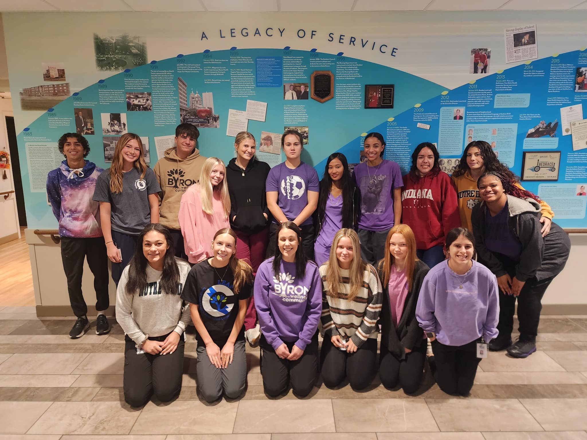 Group of young students standing and kneeling in front of a large educational display about the history of a community. The students are smiling and wearing casual clothes, some with purple hoodies and sport shirts. The display behind them features a