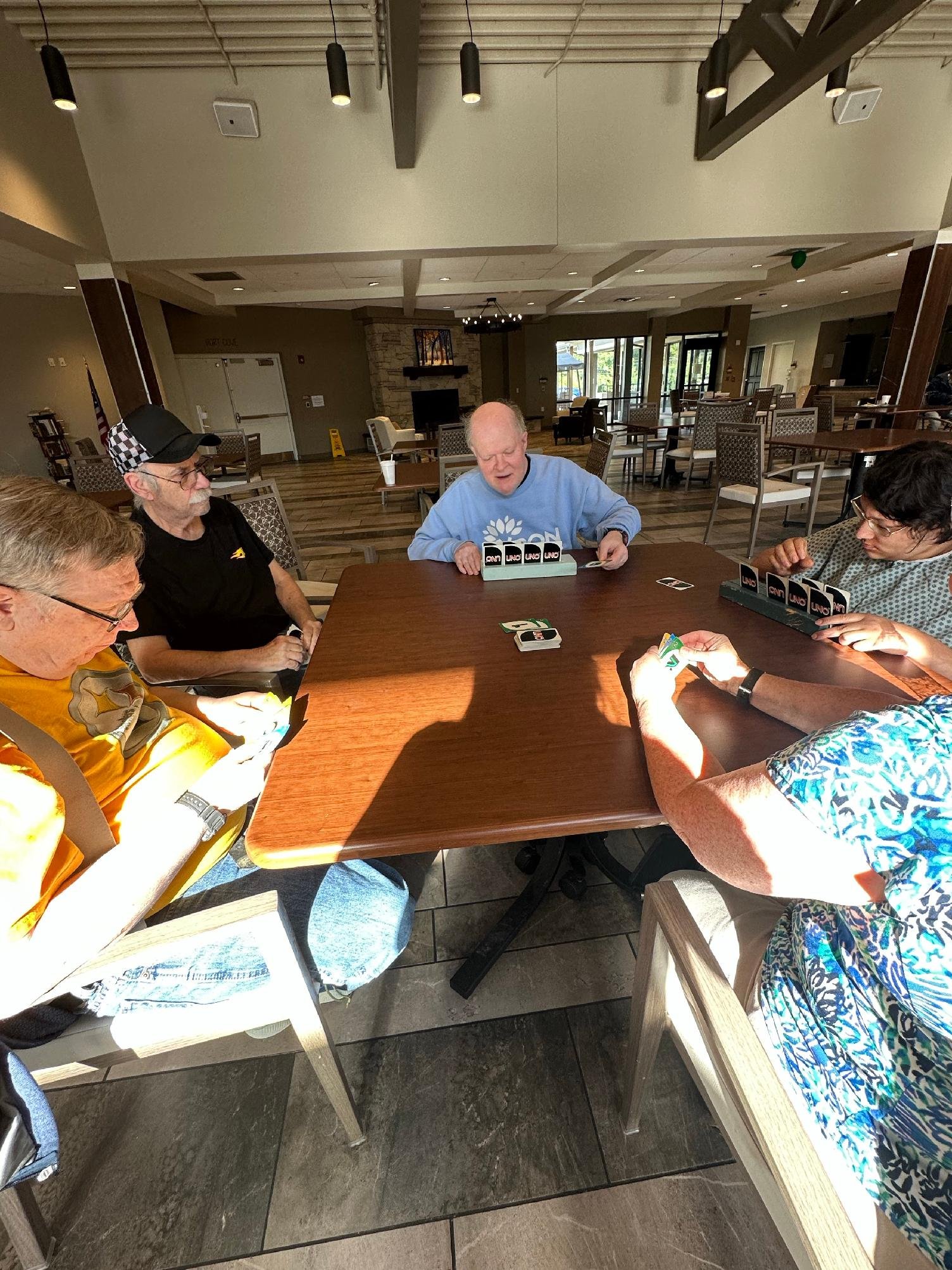 A group of people playing Uno card game at a wooden table in a spacious, well-lit indoor setting, possibly a restaurant or community area, with chairs and tables in the background.