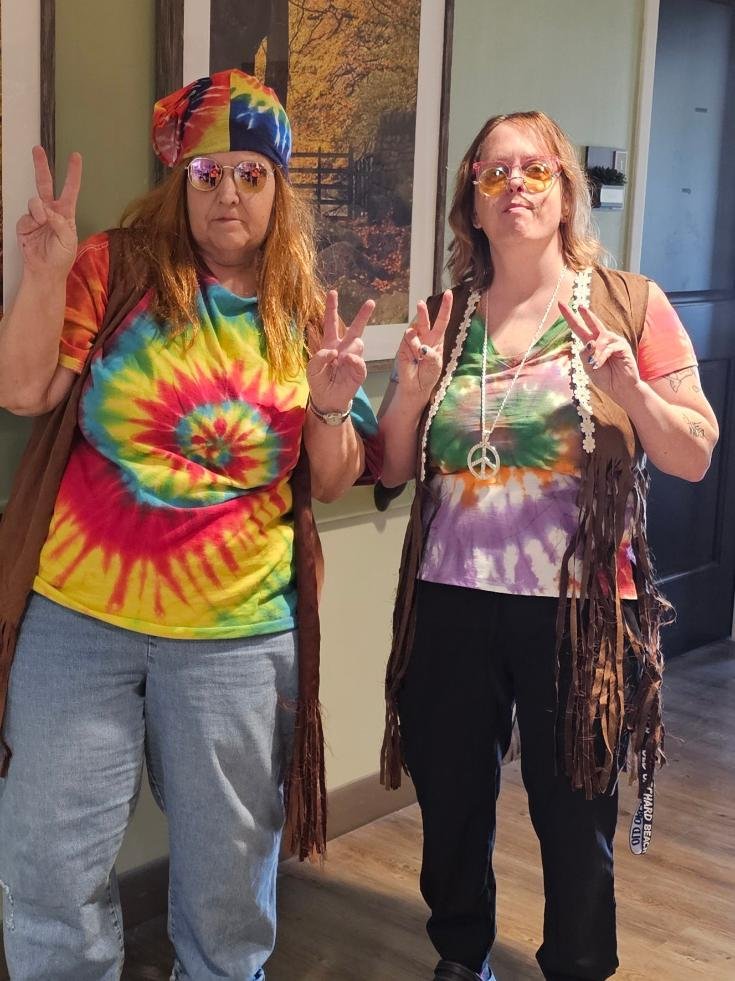 Two women dressed in colorful, hippie-style clothing making peace signs and wearing tie-dye shirts and round, retro sunglasses inside a room.
