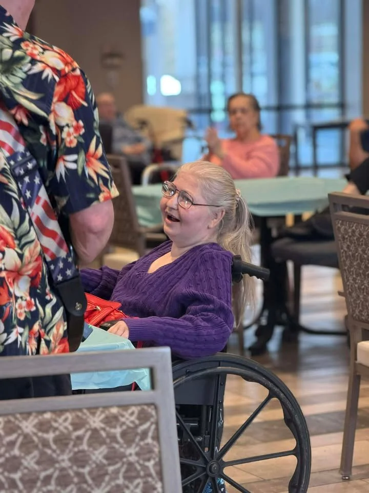 A young girl in a purple sweater and glasses smiling and engaging in conversation with a man wearing a Hawaiian shirt in a cheerful, social setting, possibly a care facility or restaurant.
