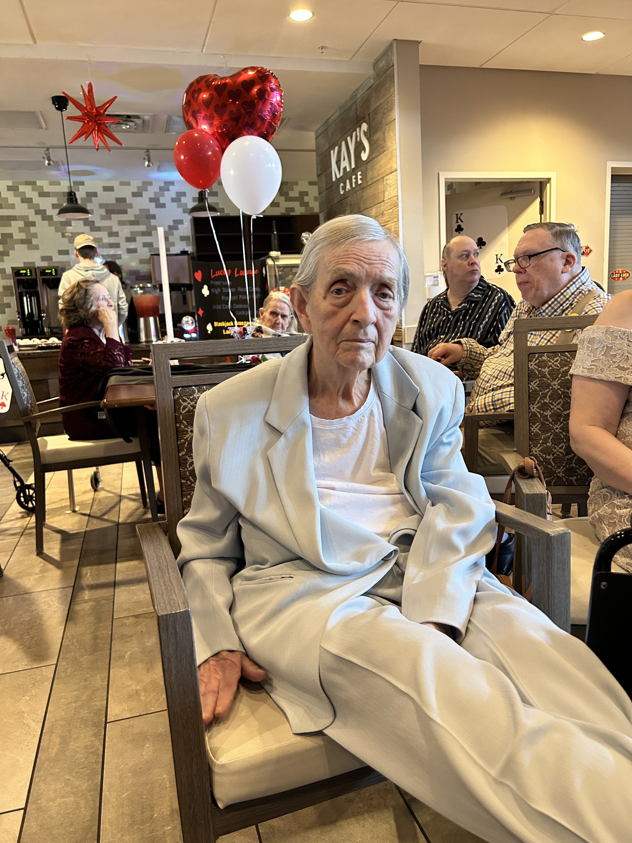 An elderly man in a gray blazer and white shirt sitting in a wooden chair at a cafe, with balloons in red, white, and a large heart-shaped red metallic balloon behind him. Several other people are visible in the background, including a woman with gra