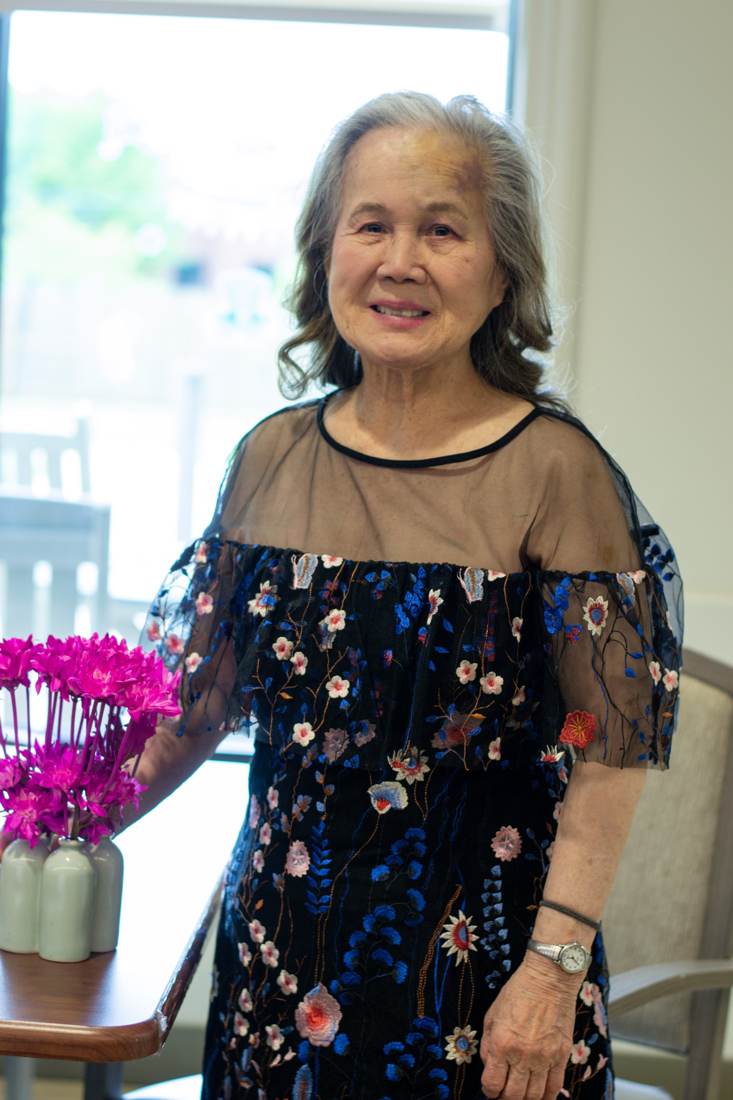 An elderly woman with gray hair standing inside near a window, wearing a black dress with colorful floral embroidery and a sheer top. She is next to a table with pink flowers in white vases and has a wristwatch on her left wrist.