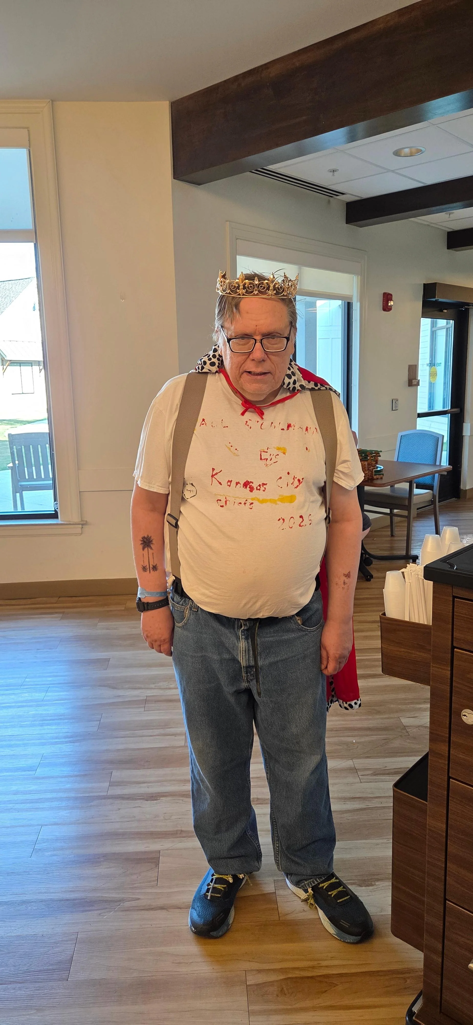 A man wearing a crown, glasses, a white t-shirt with red writing, jeans, and sneakers standing in a room with wooden floors and large windows.