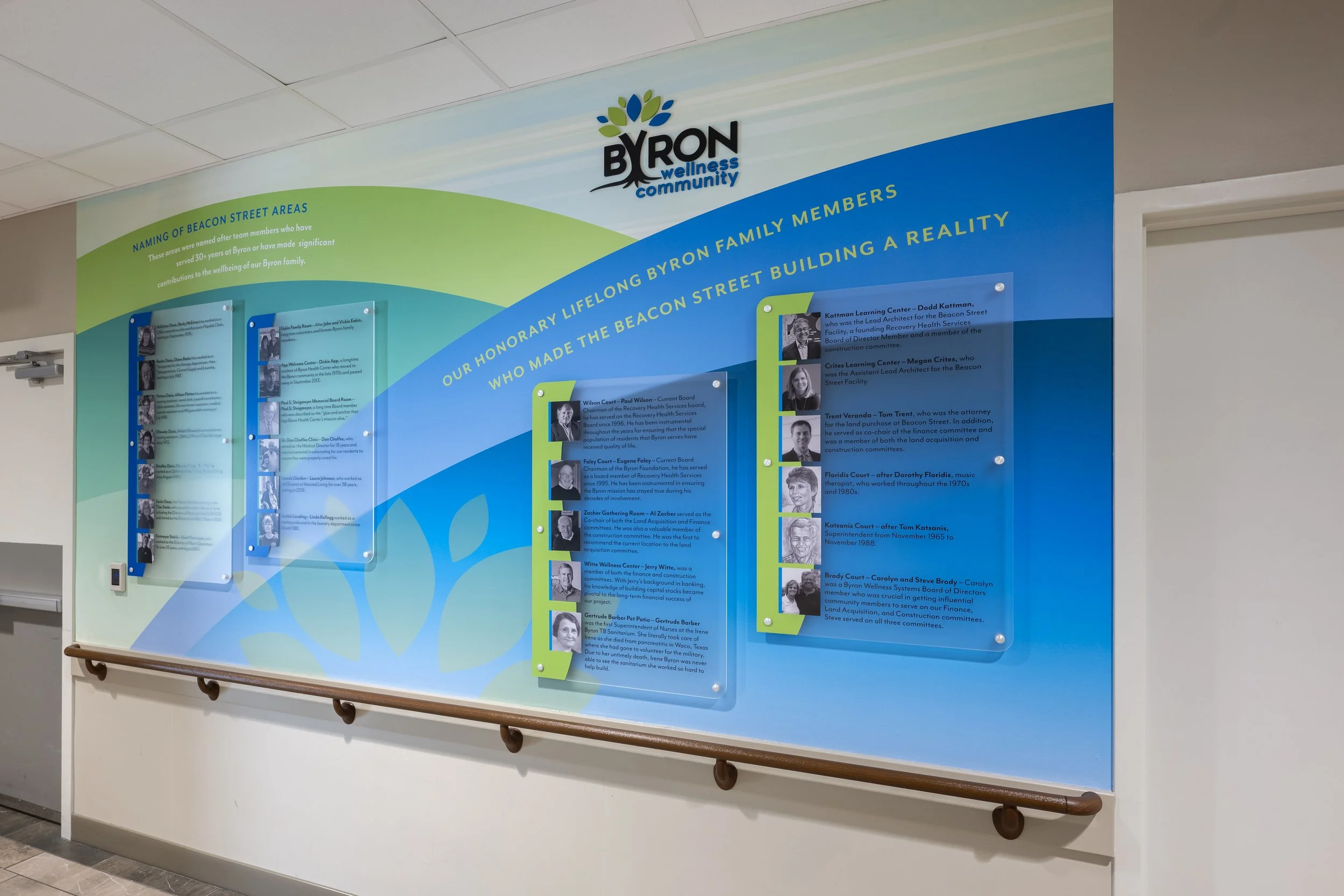A colorful wall display with the title 'Byron Wellness Community' and sections describing the history of Beacon Street areas and honoring long-time family members associated with Byron. The display includes photos of individuals along with their names and descriptions of their contributions.