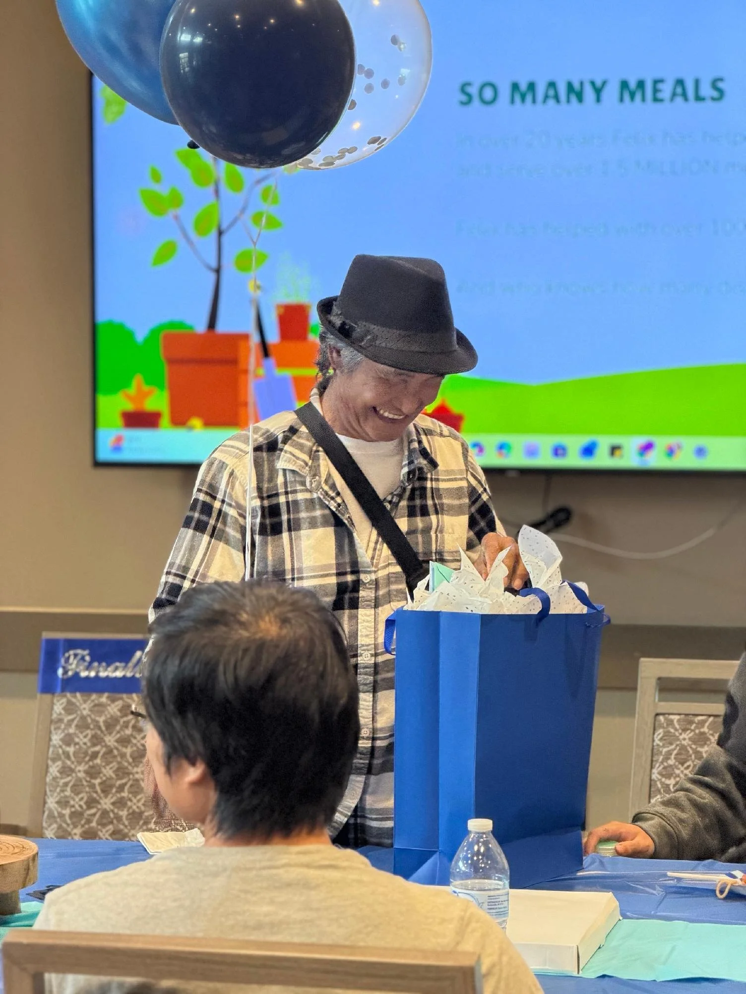 A man wearing a gray fedora and plaid shirt is smiling while opening a blue gift bag at a party or event, with balloons and a presentation screen in the background.