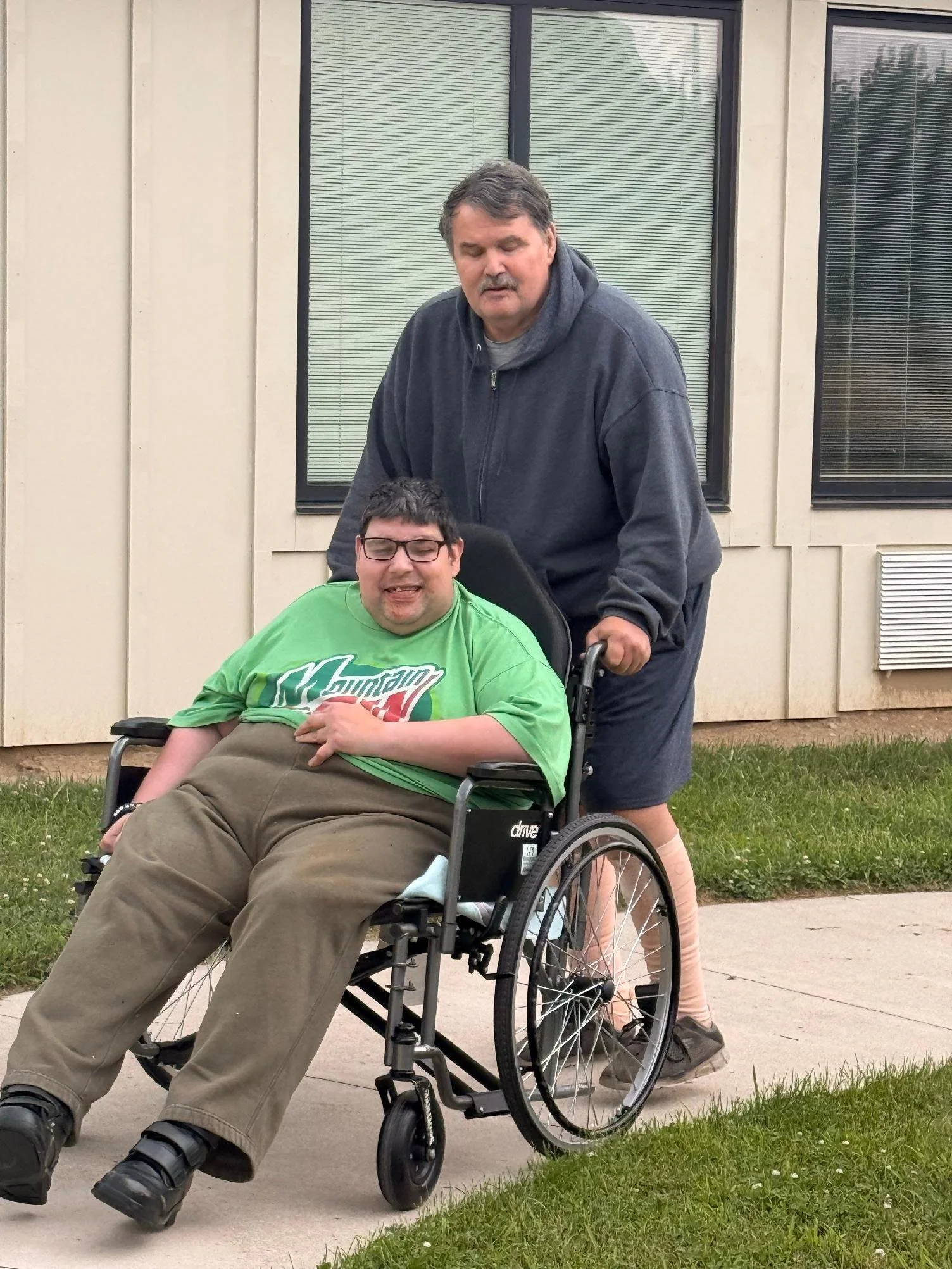 A man in a dark hoodie is standing behind a young man in a wheelchair, outside a beige building with windows. The young man is wearing glasses and a bright green Mountain Dew T-shirt, smiling with his eyes closed.