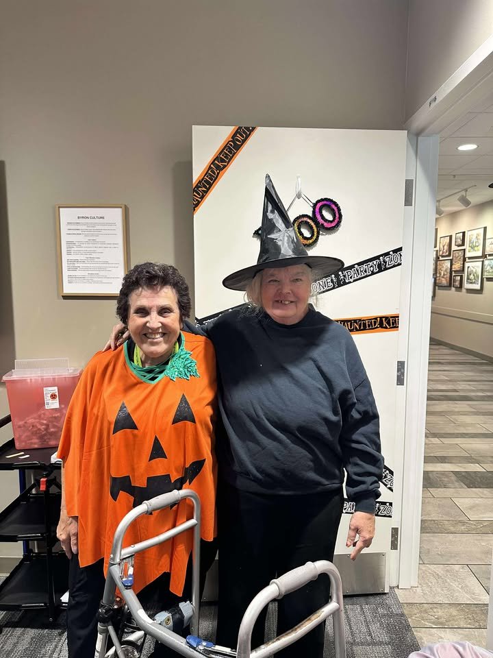 Two women dressed in Halloween costumes, one as a pumpkin with a black jack-o'-lantern face on their orange cape, and the other as a witch with a pointed hat, smiling for a photo in a hallway or lobby area.