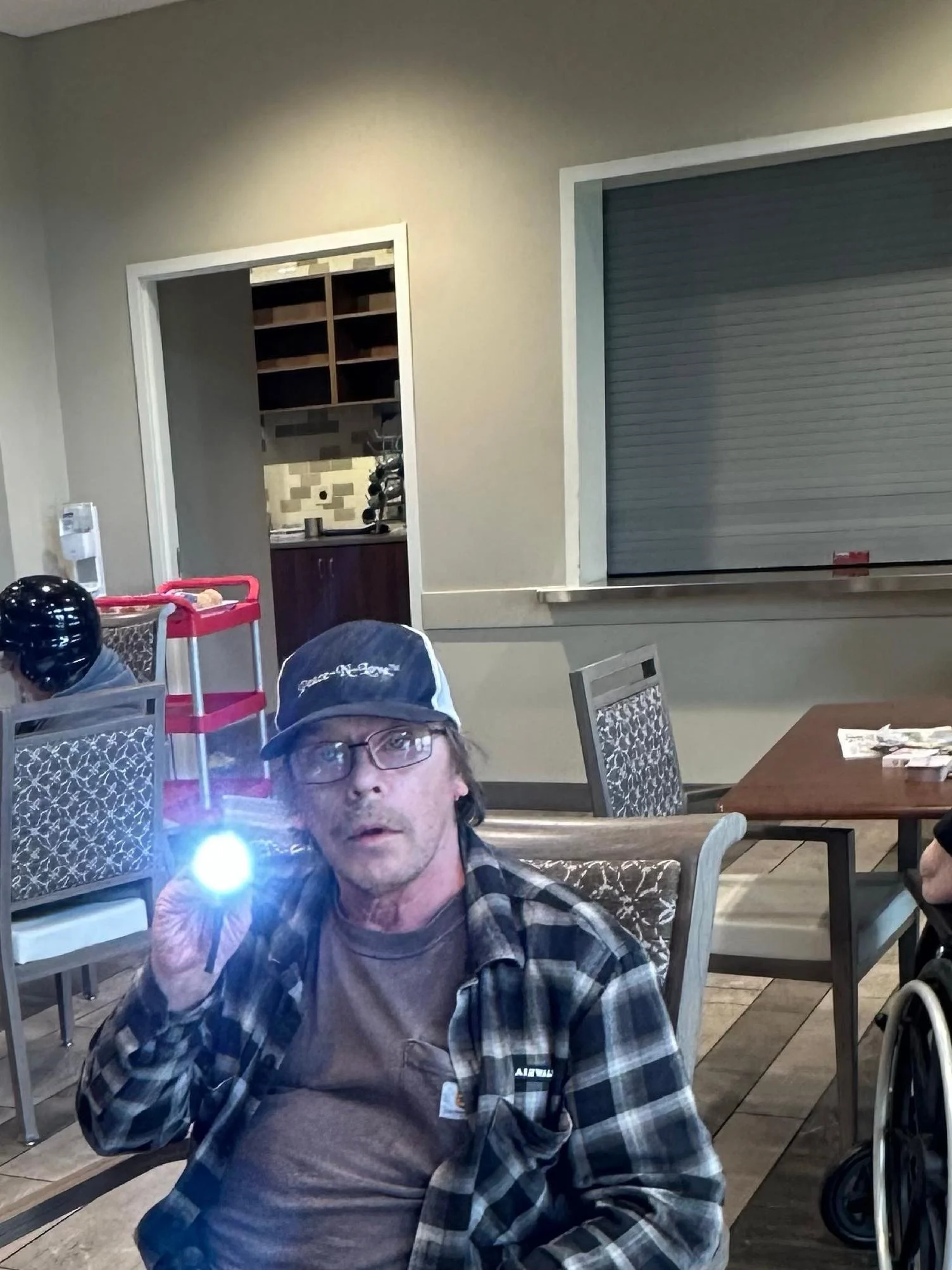 A man wearing glasses and a plaid shirt sitting in a restaurant, taking a selfie with his phone. There are chairs, tables, and a man in a wheelchair visible in the background. A rolled-down metal shutter is seen on the right side.