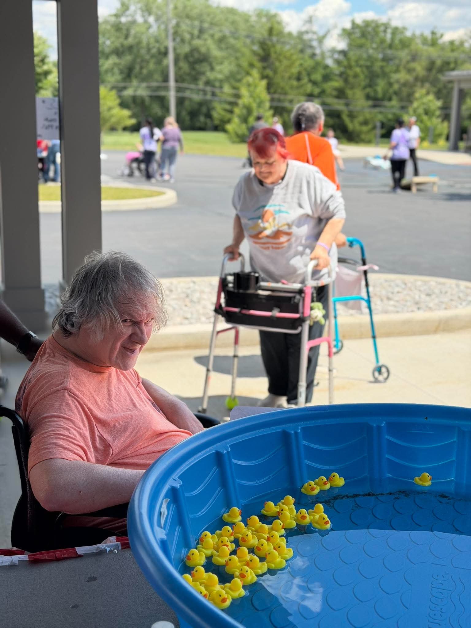 An elderly woman with gray hair and a pink shirt is sitting next to a large blue water basin filled with floating yellow rubber duck toys, outside on a sunny day. In the background, other seniors and caregivers are walking and socializing near some t