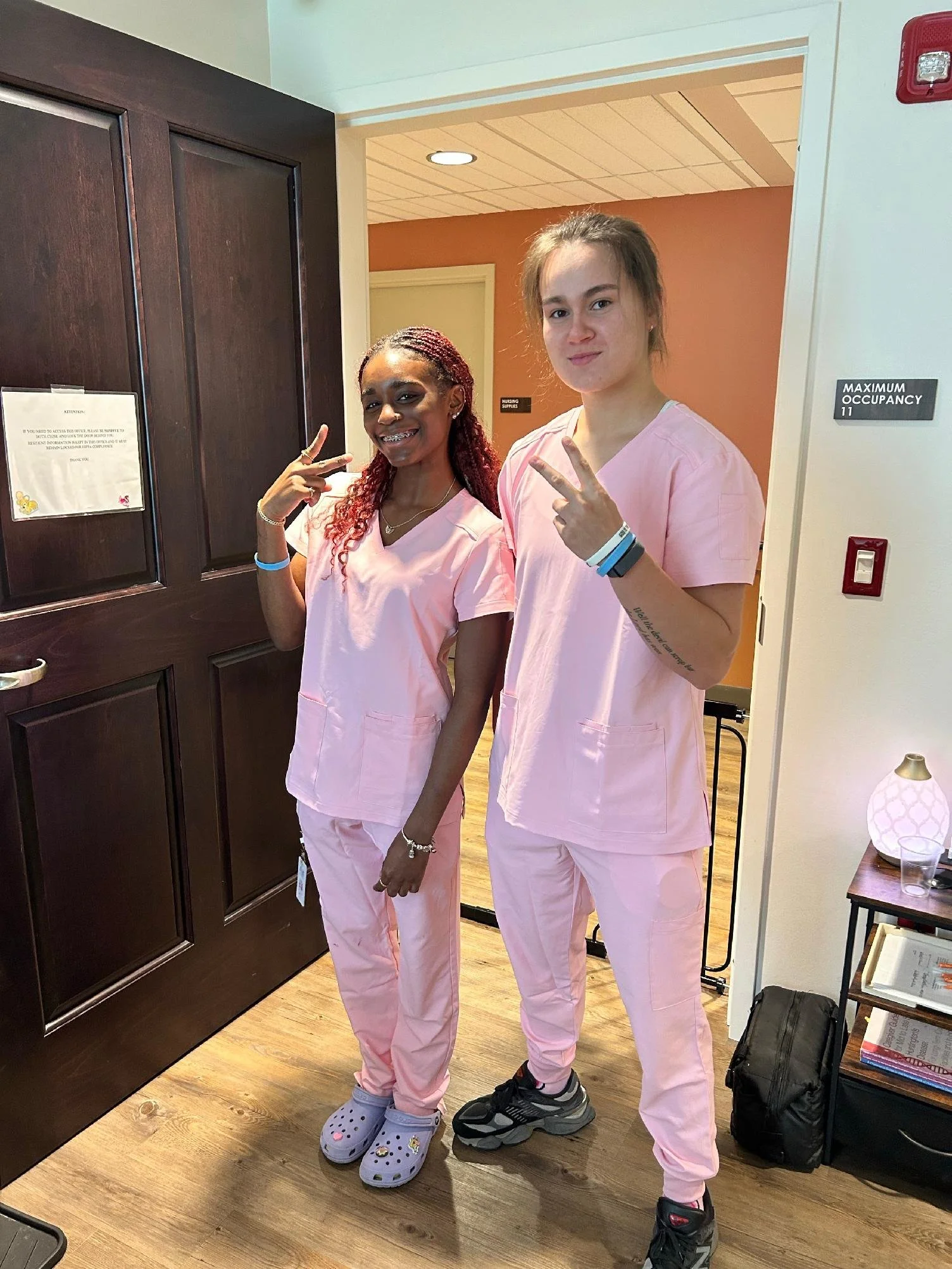 Two young women wearing pink scrubs standing in a medical or hospital setting, smiling and making peace signs with their hands.