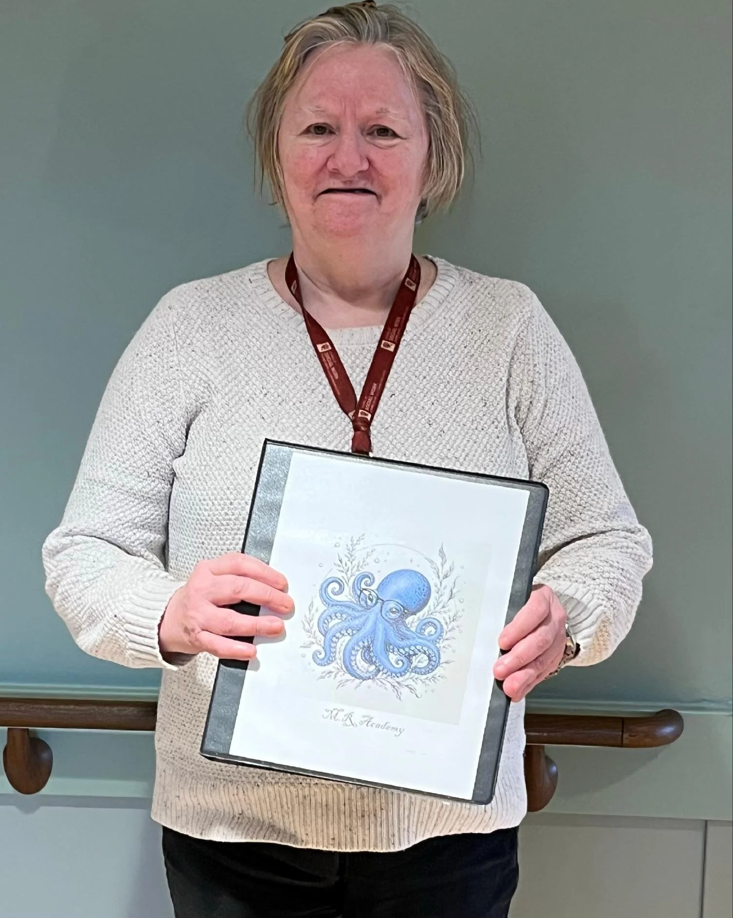 An older woman with short blonde hair wearing a cream sweater and a maroon lanyard, holding a framed artwork of a blue octopus with decorative elements, standing in front of a gray wall.