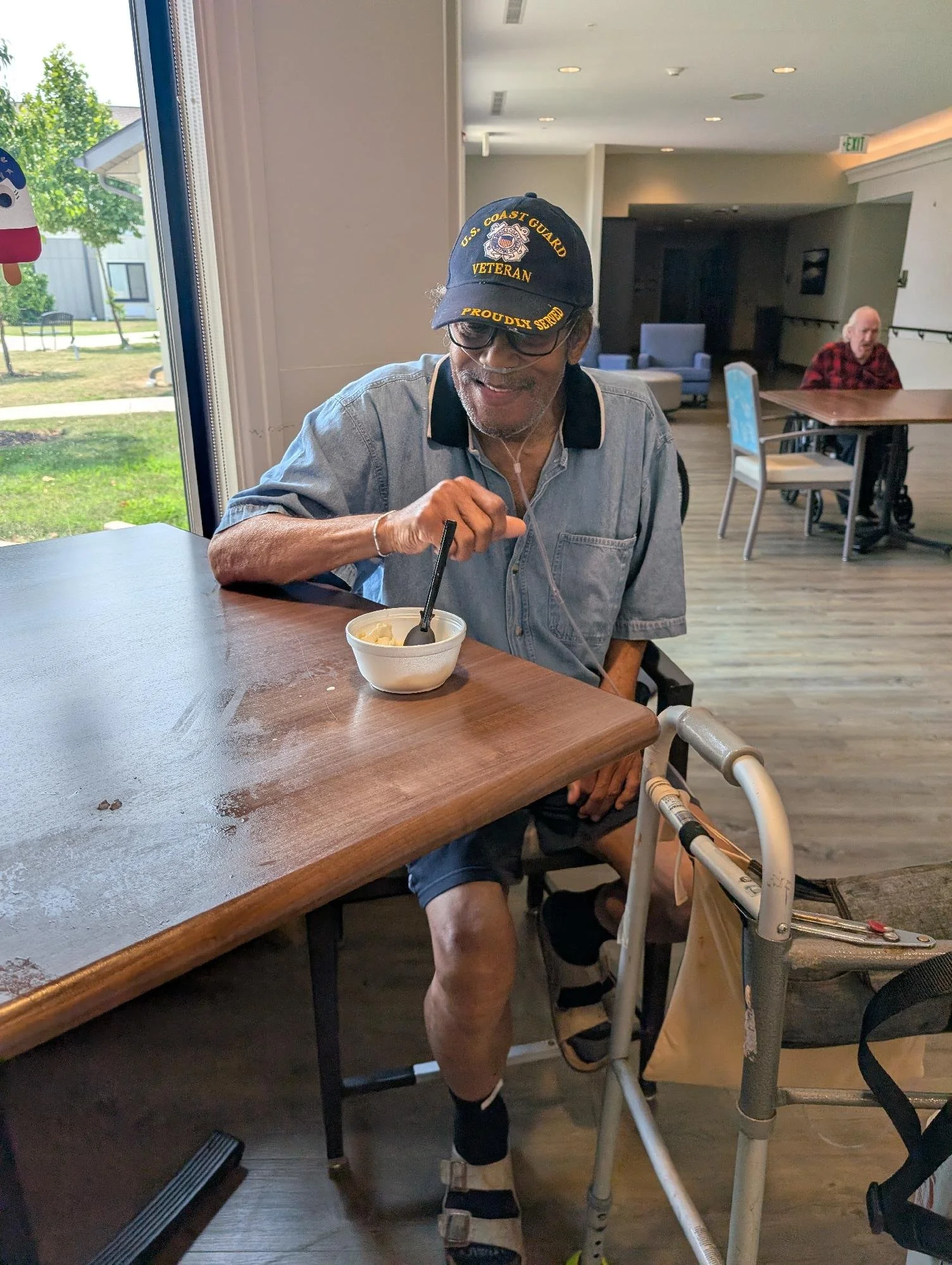 A smiling elderly man with glasses, wearing a U.S. Coast Guard veteran cap, sitting at a table with a bowl of ice cream, using a walker nearby, inside a well-lit room.