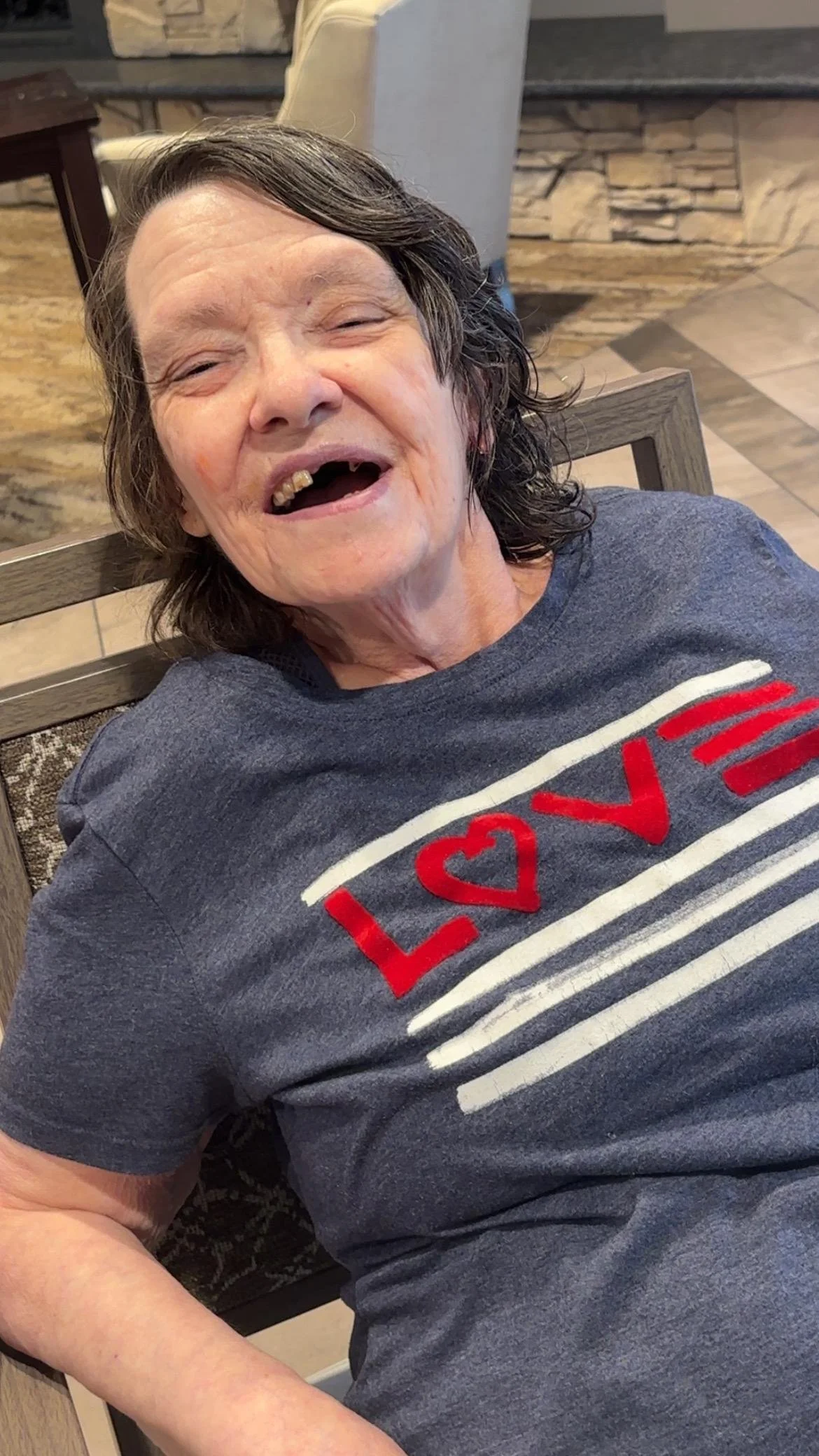 A woman with short dark hair smiling and showing missing teeth, wearing a gray T-shirt with a red, white, and black design, sitting in a restaurant.