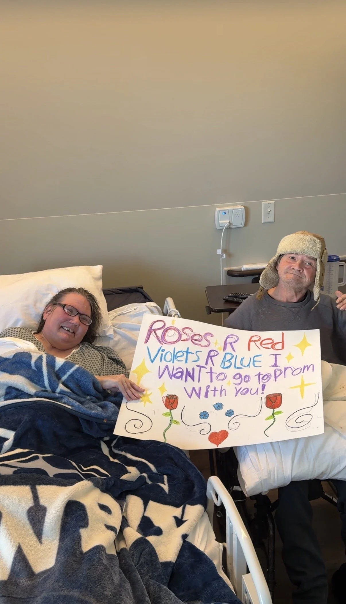 A woman lying in a hospital bed holding a colorful handmade sign that says 'Roses R Red, Violets R Blue I Want to go to prom with you!' Next to her, a man wearing a beige trapper hat and gray shirt is sitting in a wheelchair, smiling at the camera.