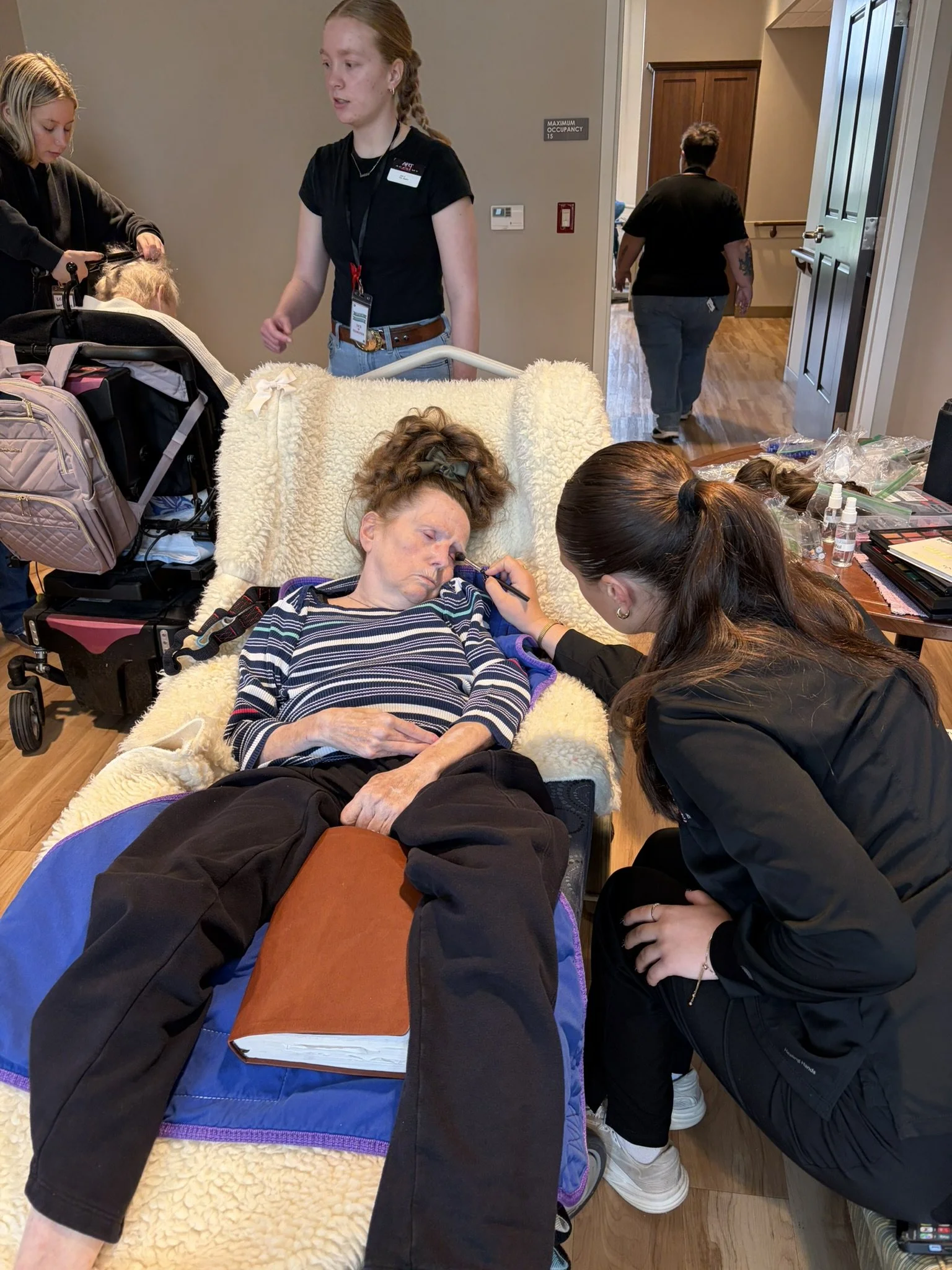 An elderly woman laying in a hospital bed with her eyes closed, while a woman in black, crouching, applies makeup or face paint to her face. Other woman is sitting beside her. Medical supplies and personal items are on the bedside table. In the backg