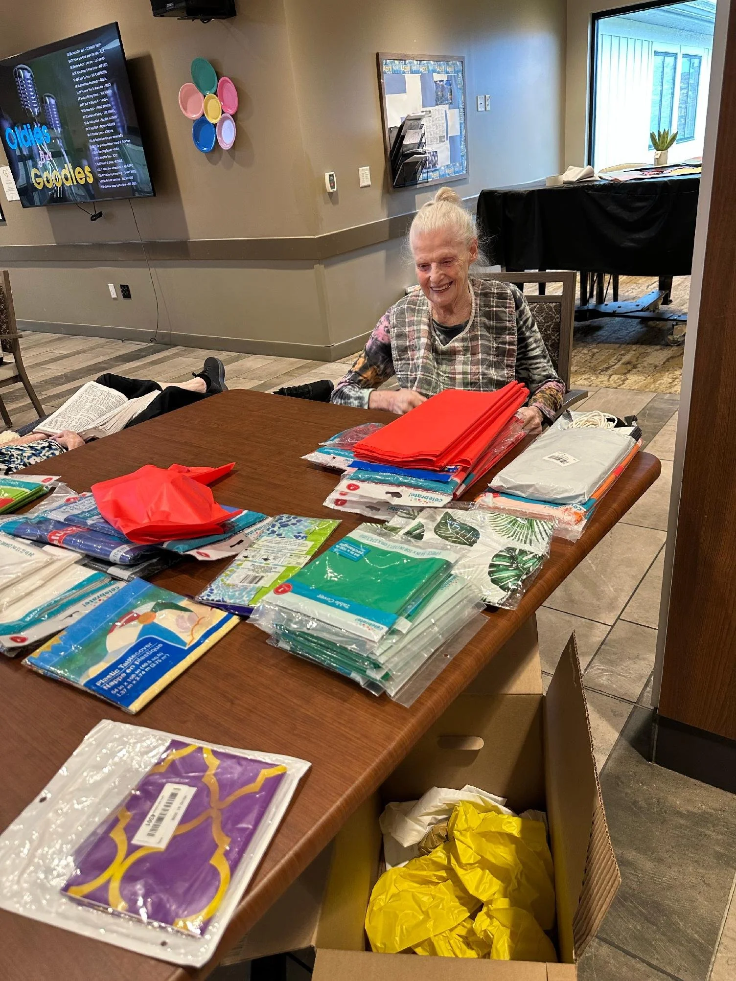 A smiling elderly woman sitting at a table with craft supplies, including colorful tissue paper, plastic zip bags, and paper napkins, in a room with beige walls, a large window, and a TV screen on the wall.