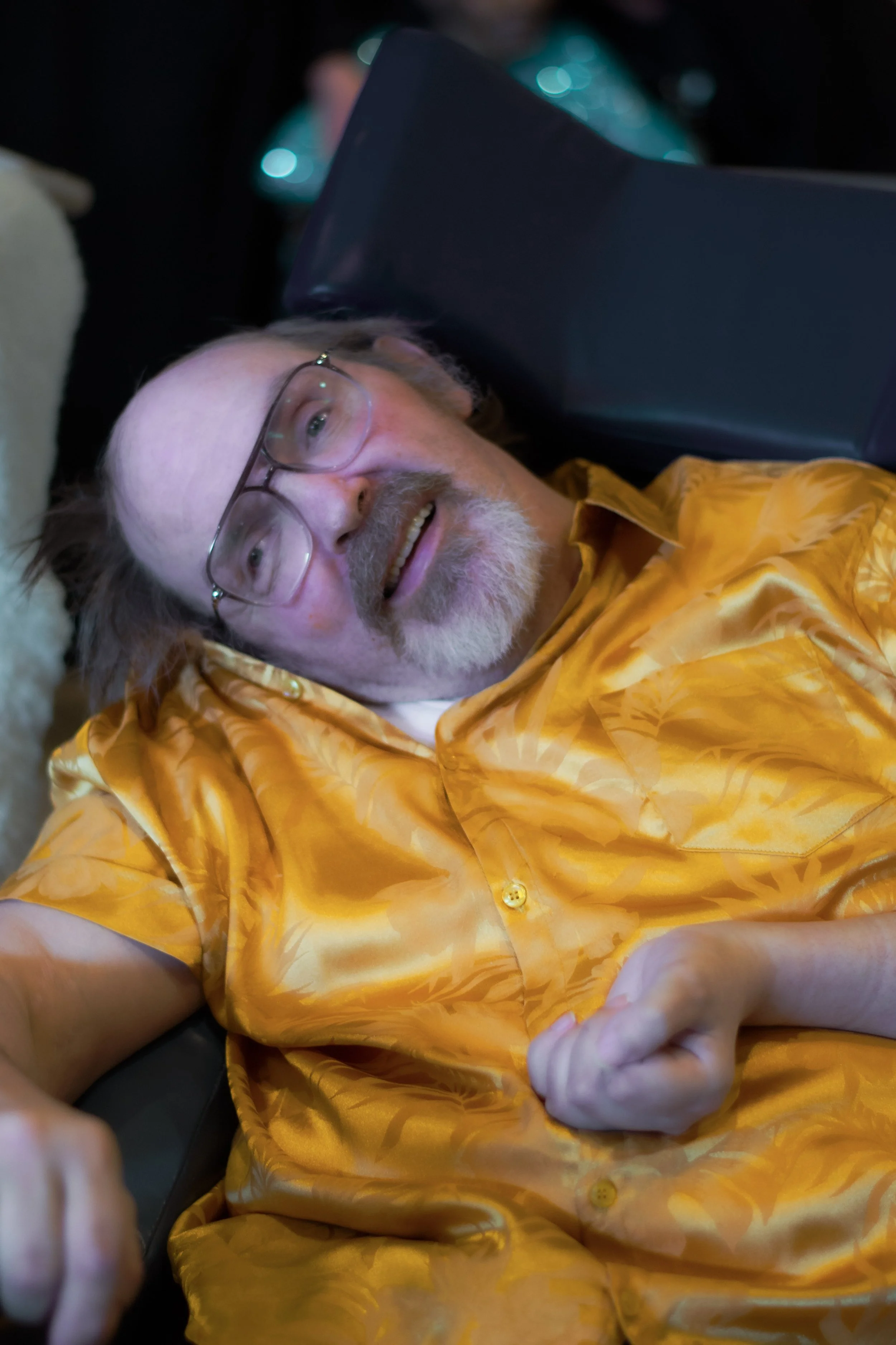 A man with glasses and a beard lying down, wearing a yellow patterned shirt, smiling and taking a selfie.