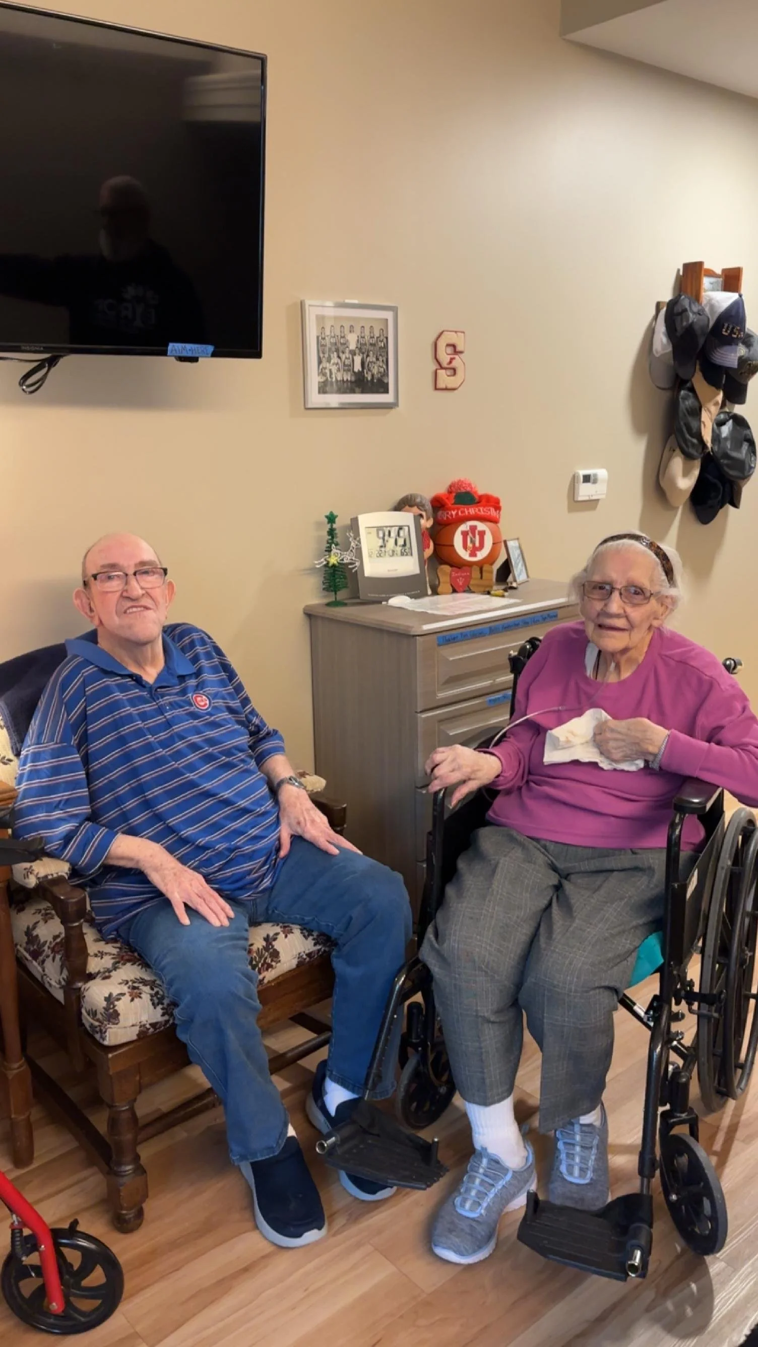 Two elderly people, one man and one woman, sitting in a room with decorations and hats, smiling. The woman is in a wheelchair.