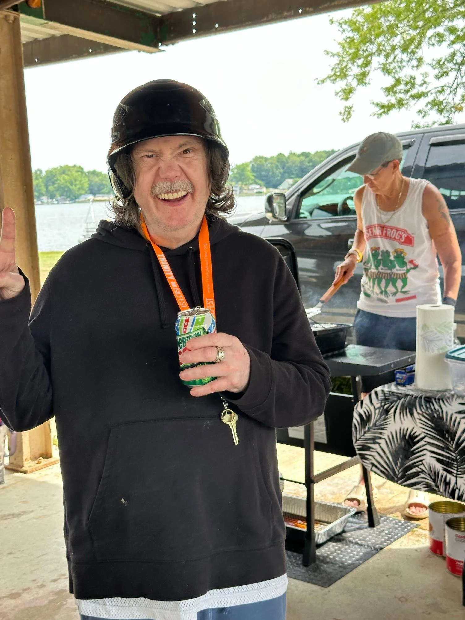 A man wearing a black motorcycle helmet, black hoodie, and holding a soda can, smiling and gesturing with his thumb. In the background, there is a woman at a grill wearing a tank top and baseball cap, cooking outdoors near a vehicle and a lake or riv