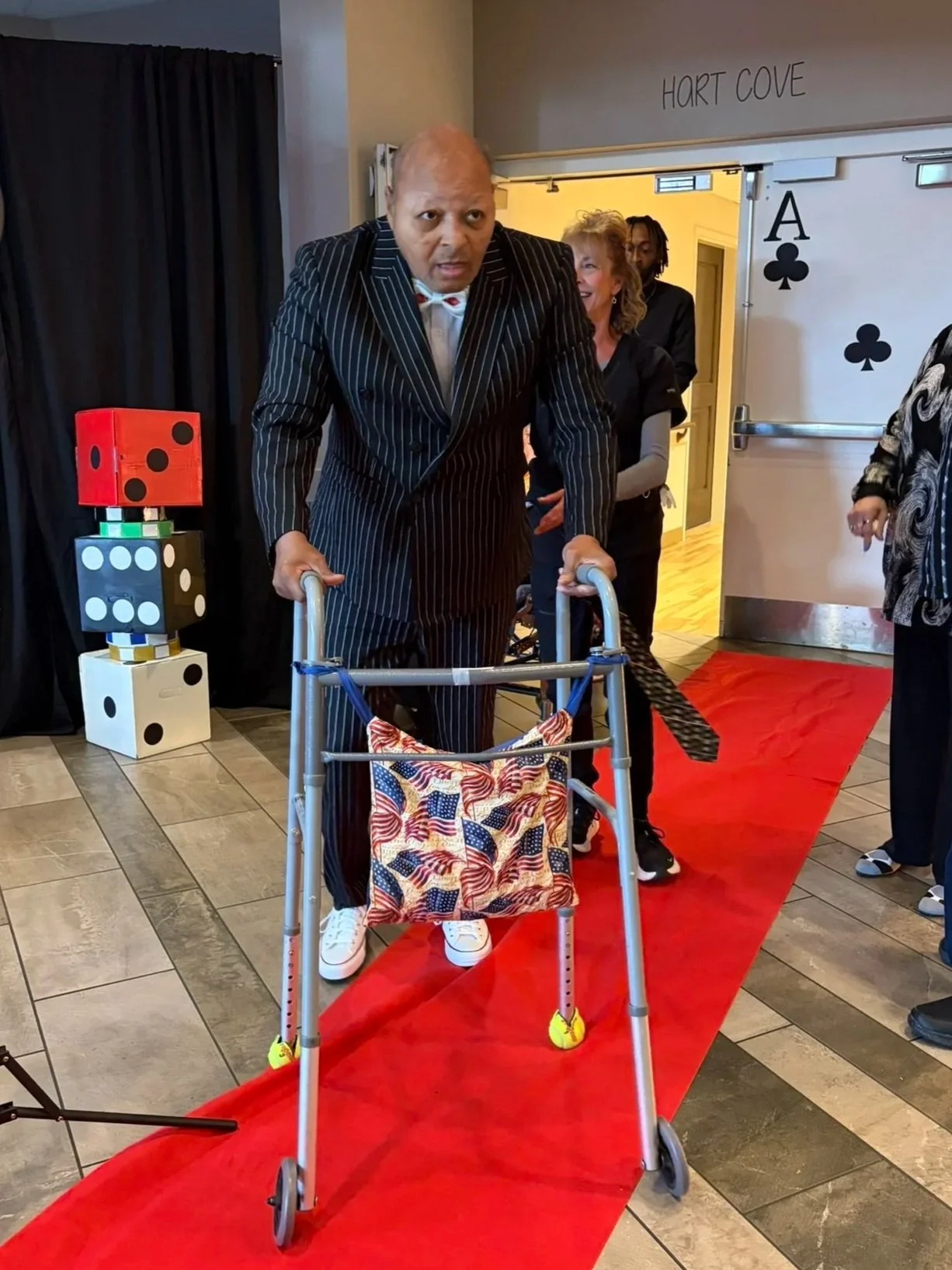 A man with a walker decorated with American flags walking on a red carpet. He is wearing a pinstriped suit with white sneakers. Behind him, a woman is smiling and other people are present in the background at an indoor event.
