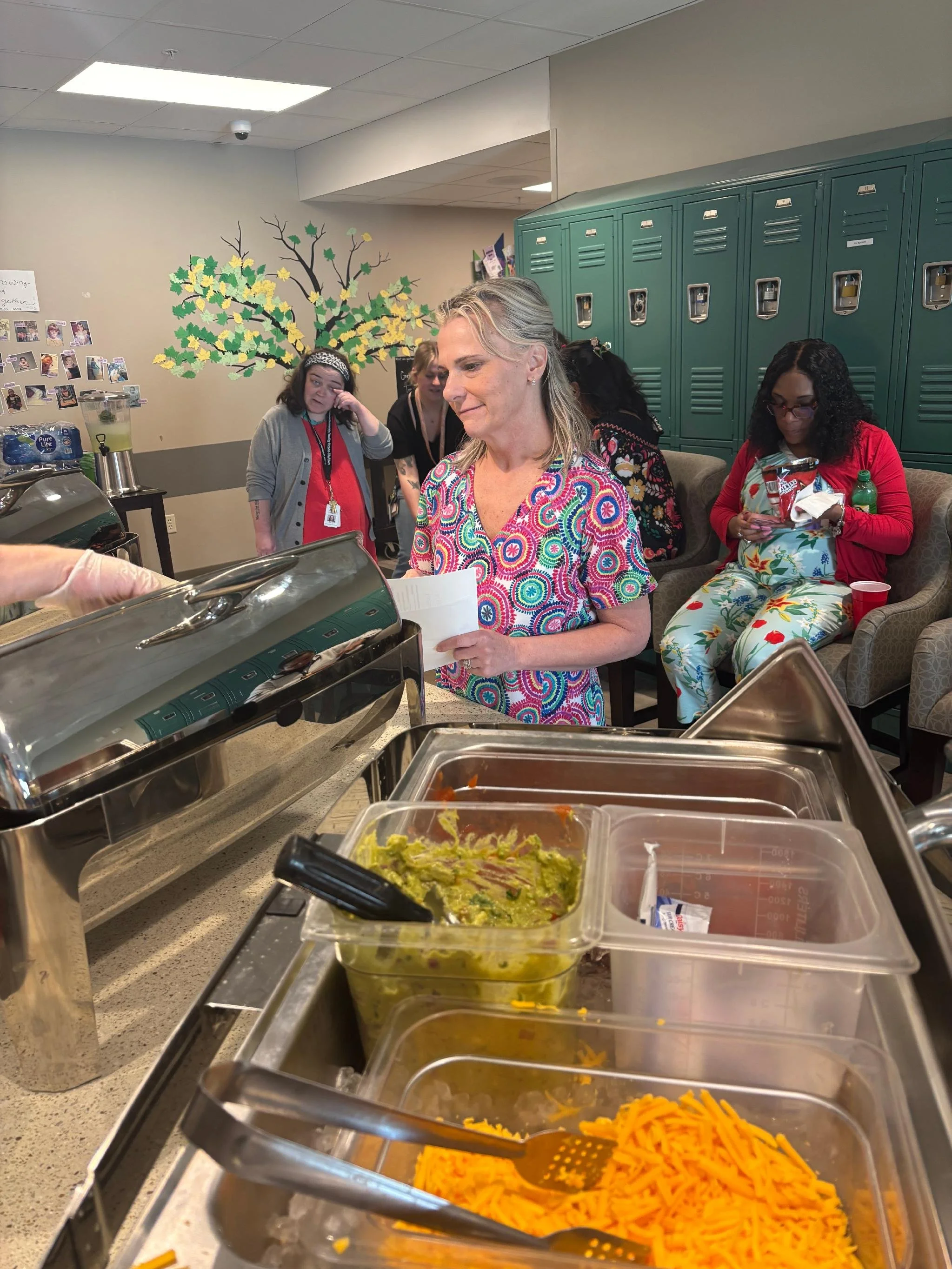 People serving food at a buffet table with containers of shredded cheese, guacamole, and other dishes. Wall with a painted tree and lockers in the background.