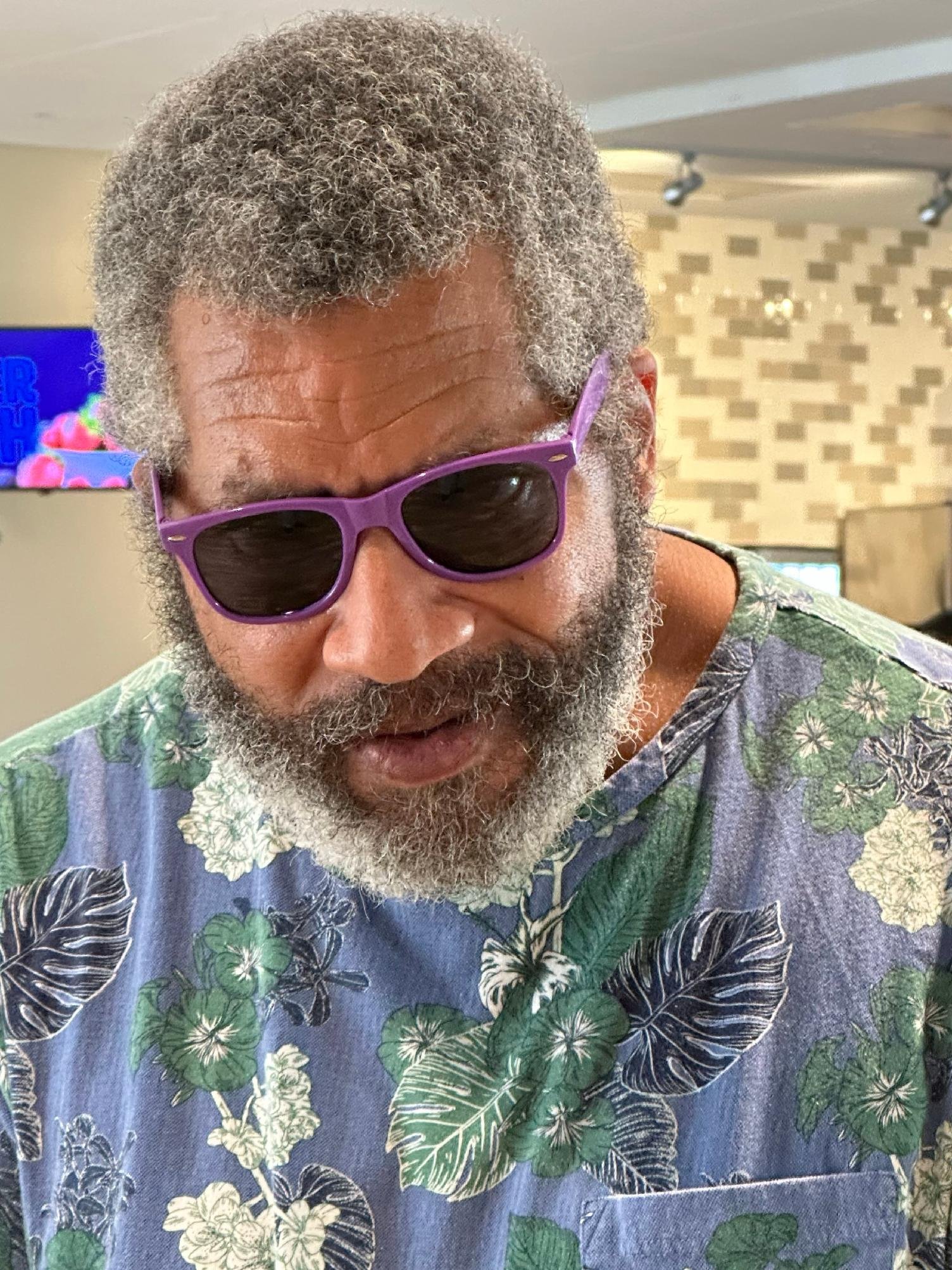 Close-up of an older man with gray curly hair and beard wearing purple sunglasses and a Hawaiian shirt with green, white, and black floral patterns, indoors with a television in the background.