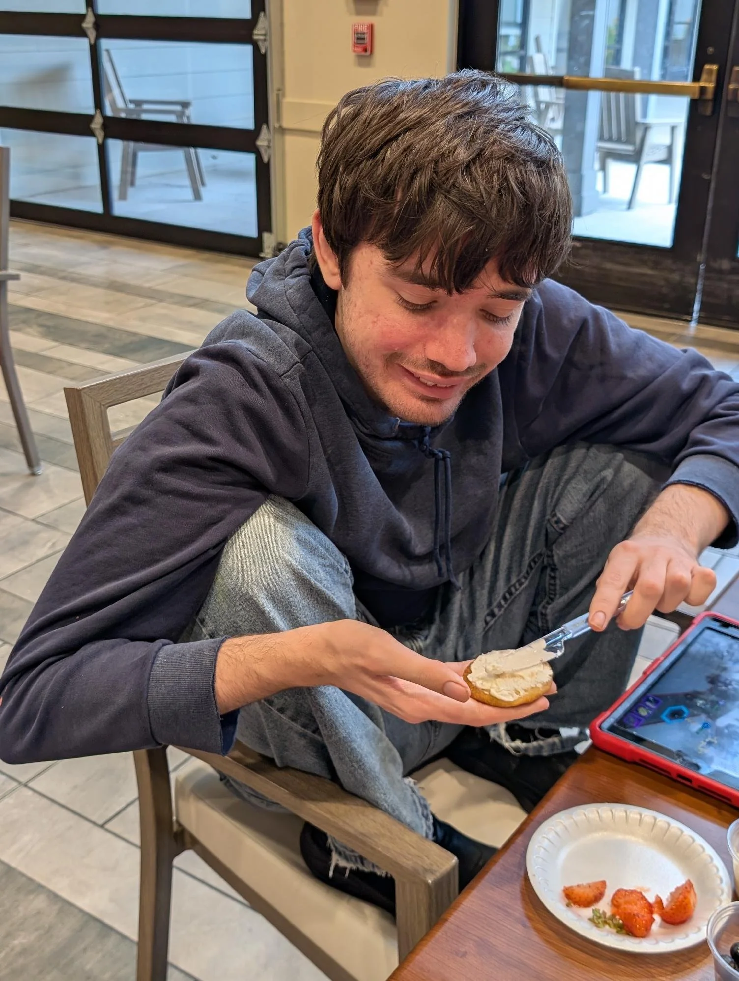 A young man with brown hair and a beard, wearing a dark hoodie and jeans, sitting in a wooden chair at a table, spreading cream cheese on a bagel. On the table, there's a white paper plate with strawberries and other fruit. Behind him by the window a