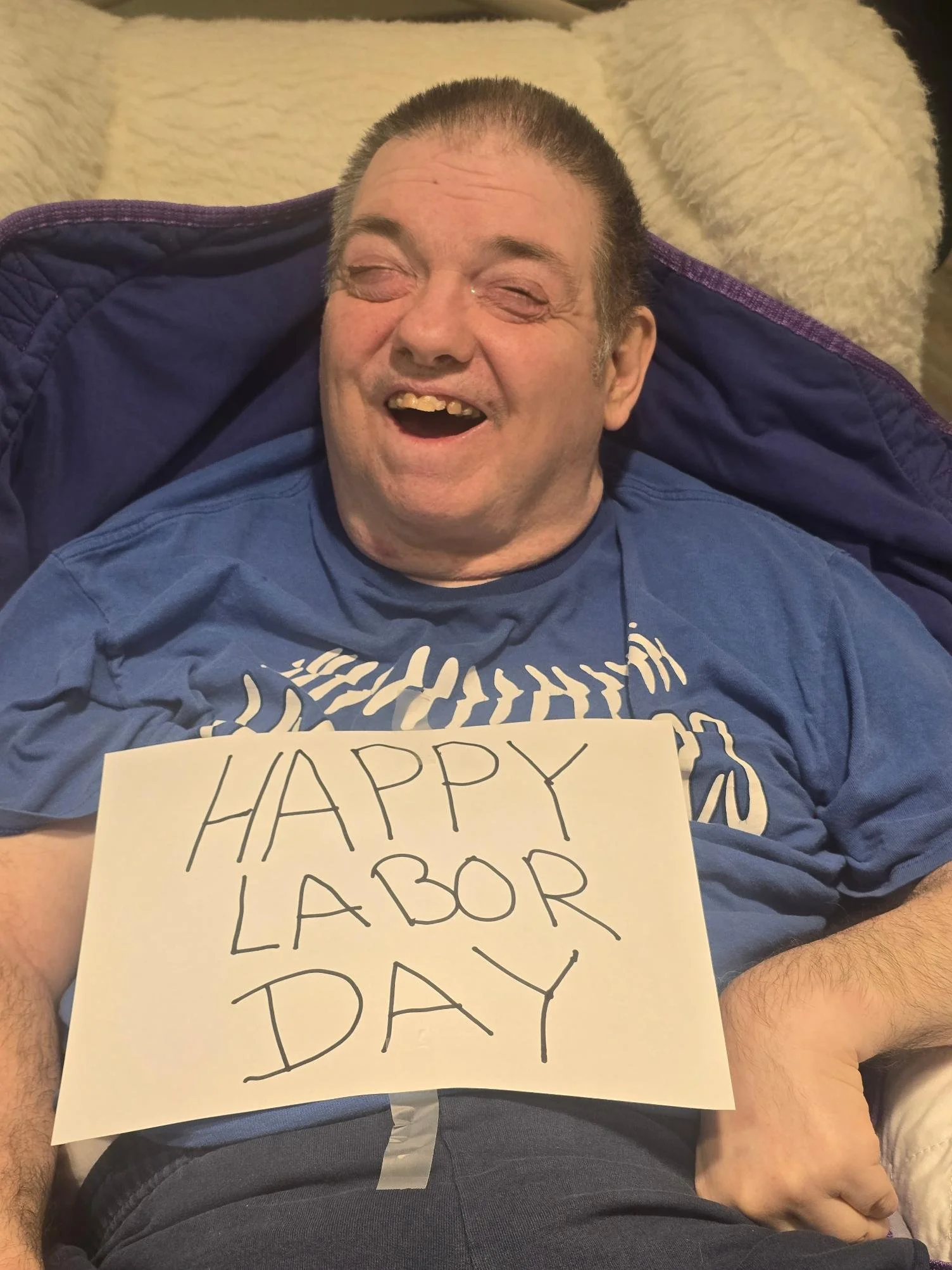 A man with short hair, smiling with his eyes closed, wearing a blue t-shirt, holding a sign that says 'HAPPY LABOR DAY', sitting against a beige fluffy blanket.